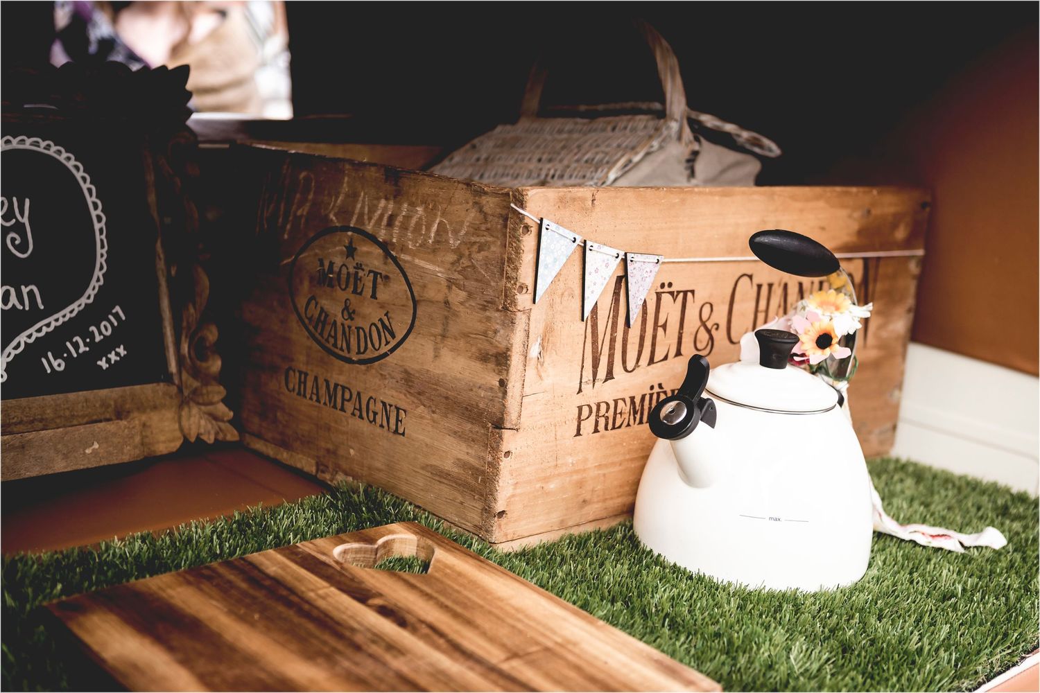 Vintage wooden crates and a white ceramic teapot arranged on artificial grass in moody lighting.