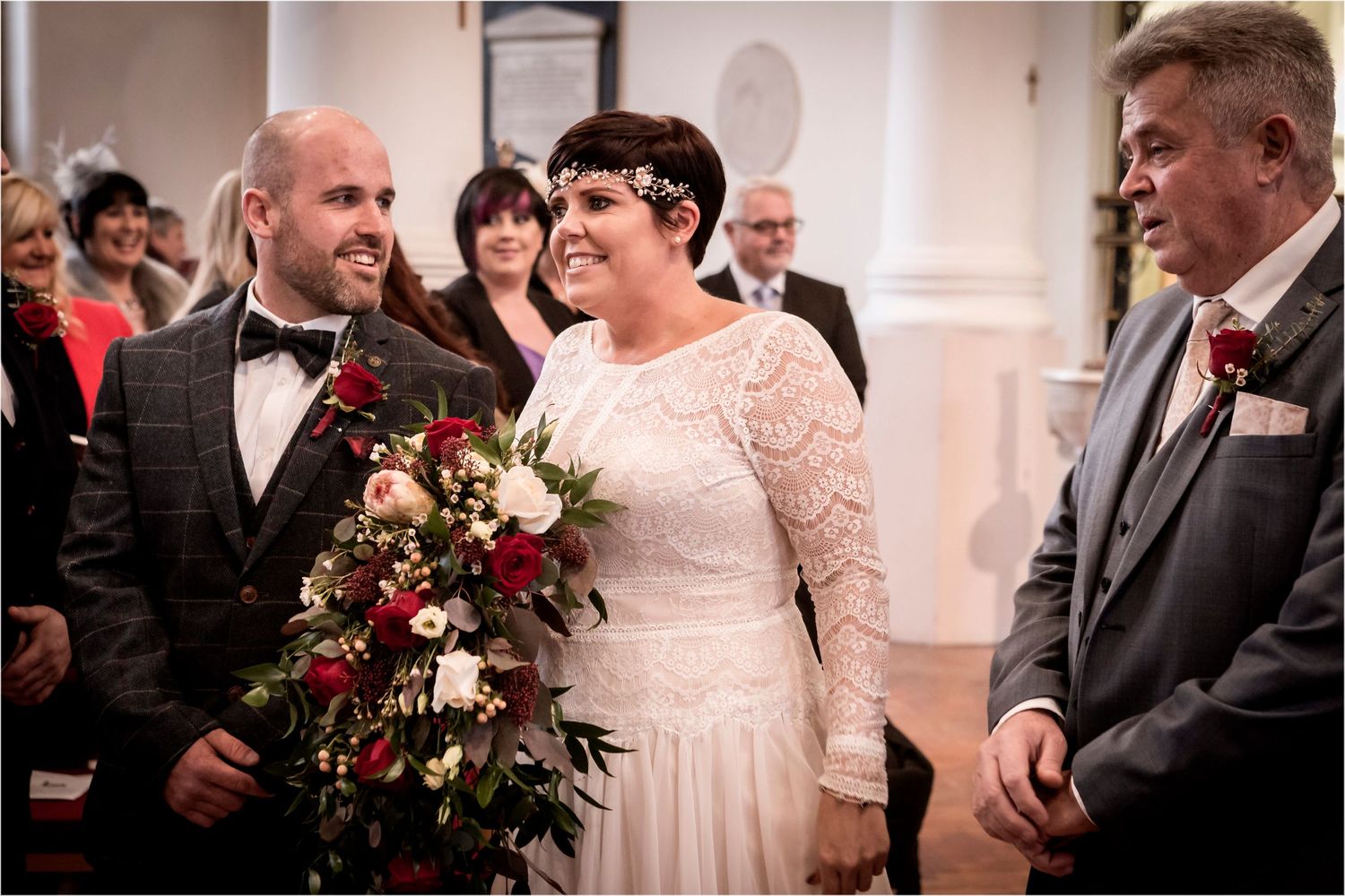 A joyful couple shares a candid moment at their wedding ceremony, with a gorgeous deep red and white floral bouquet.