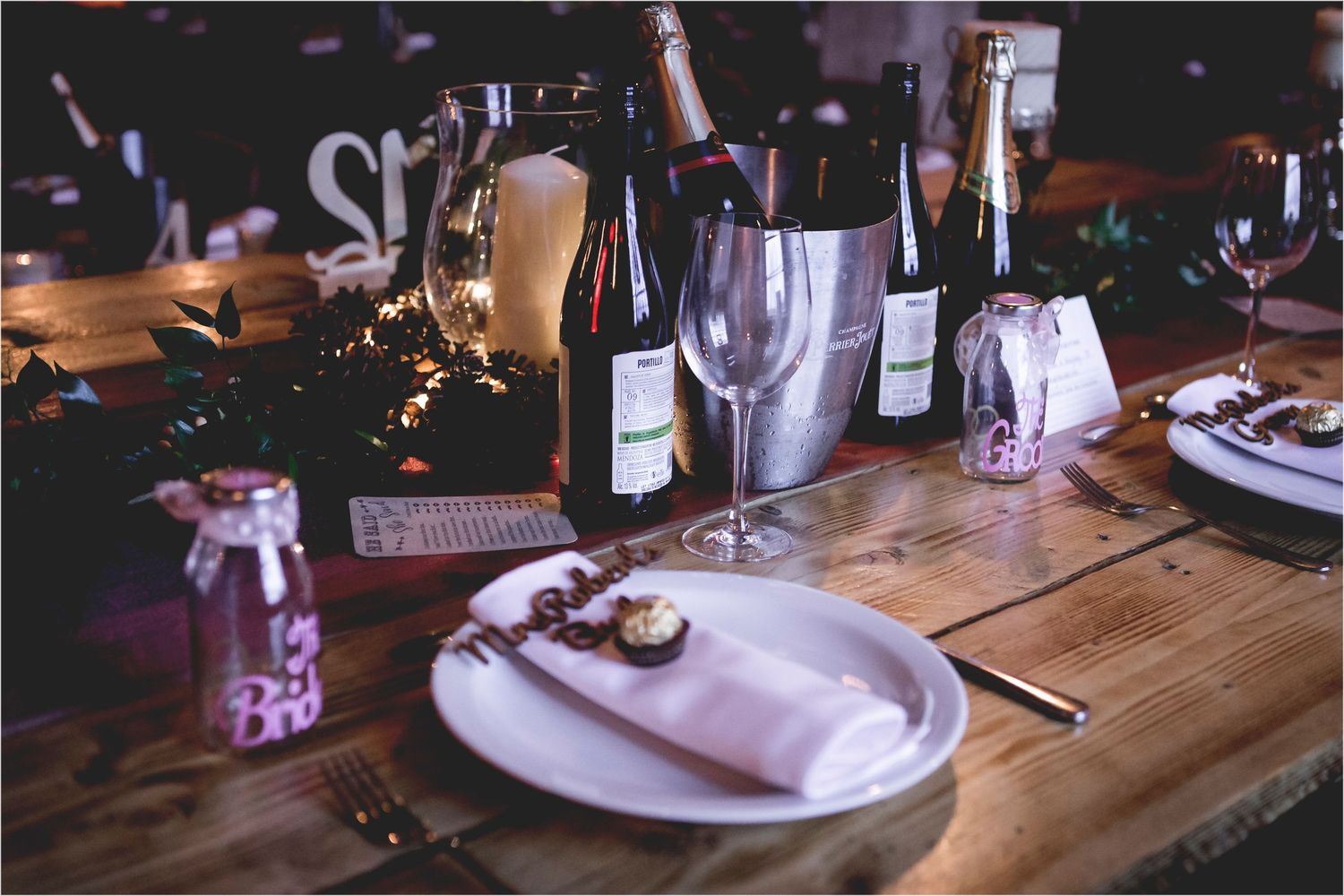 A rustic wooden table set for dinner with wine bottles, white plates, and soft candlelight for an intimate gathering.
