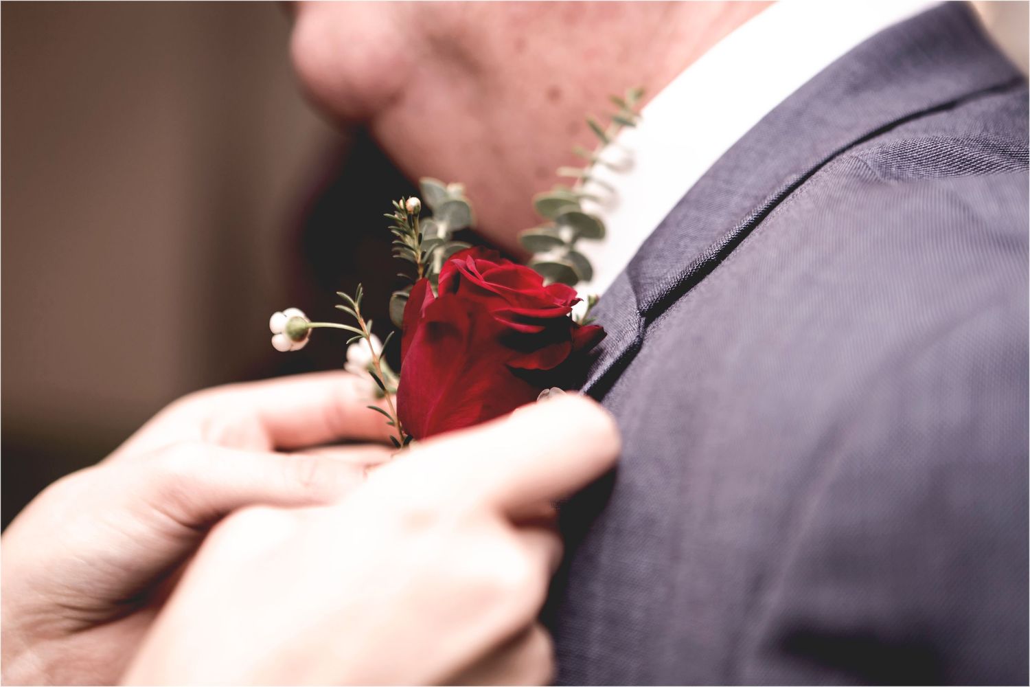 Someone pins a red rose boutonniere with small white flowers onto a grey suit lapel.