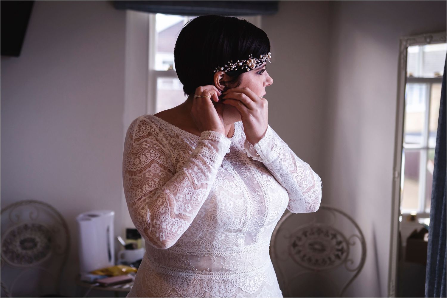 Bride in elegant white lace long sleeve wedding gown getting ready for her special day by window light.