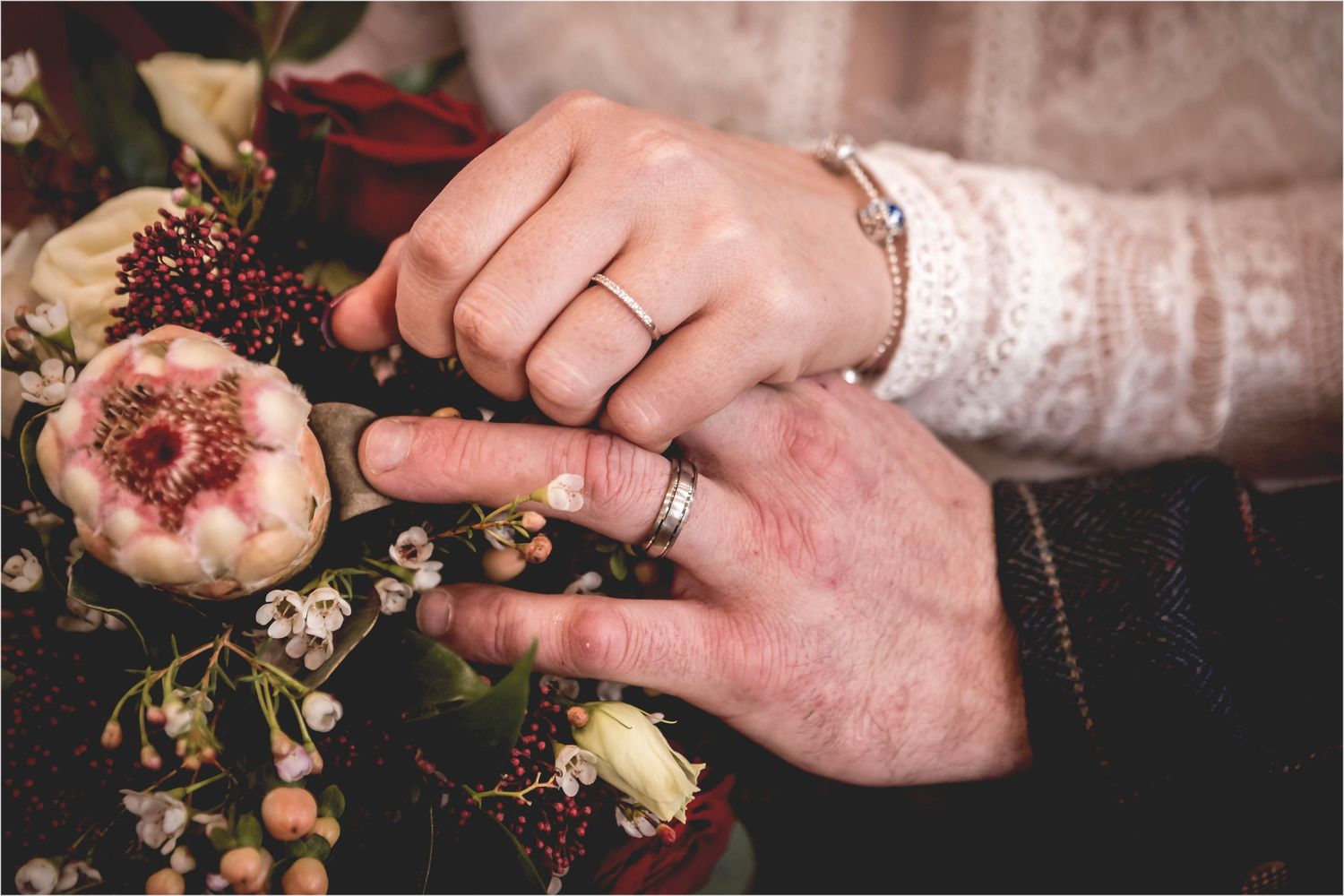 Close-up of hands showing a wedding ring with dried flowers and lace sleeves in the background.