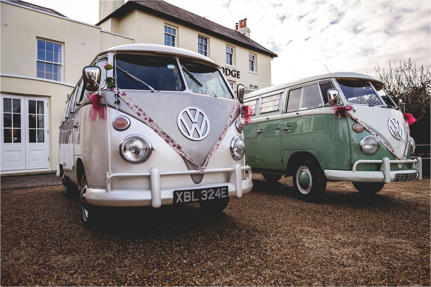 Two vintage VW split-window buses, one pink and one green, parked in front of a white house with gravel driveway.