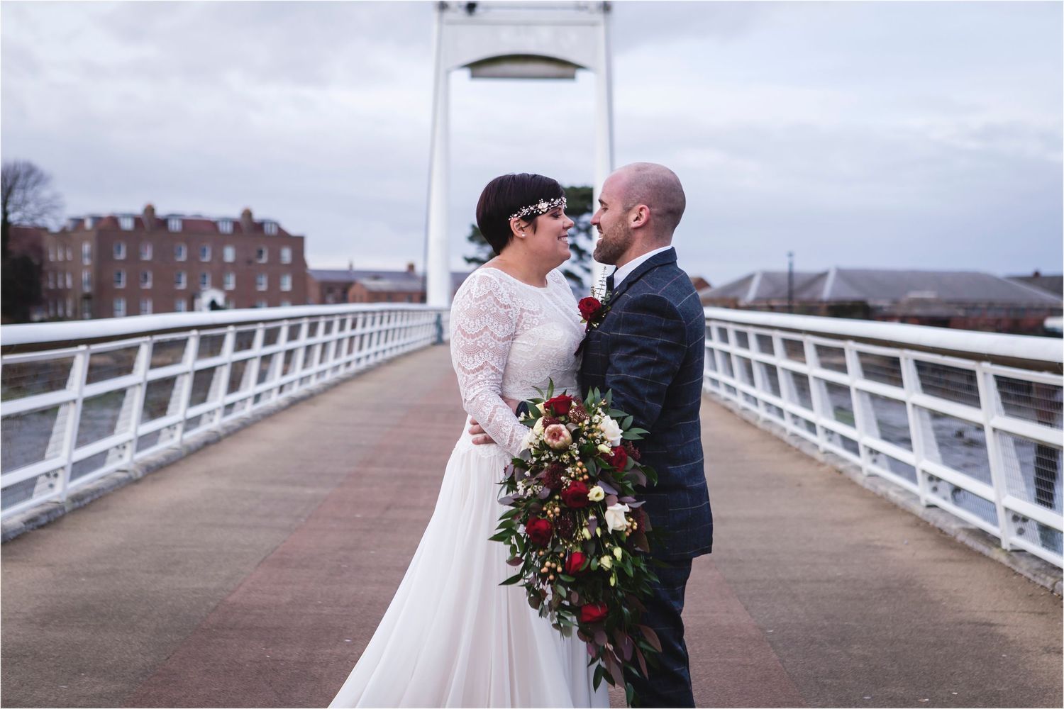 A couple shares a romantic moment on a white bridge walkway while holding a dramatic burgundy and red floral bouquet.