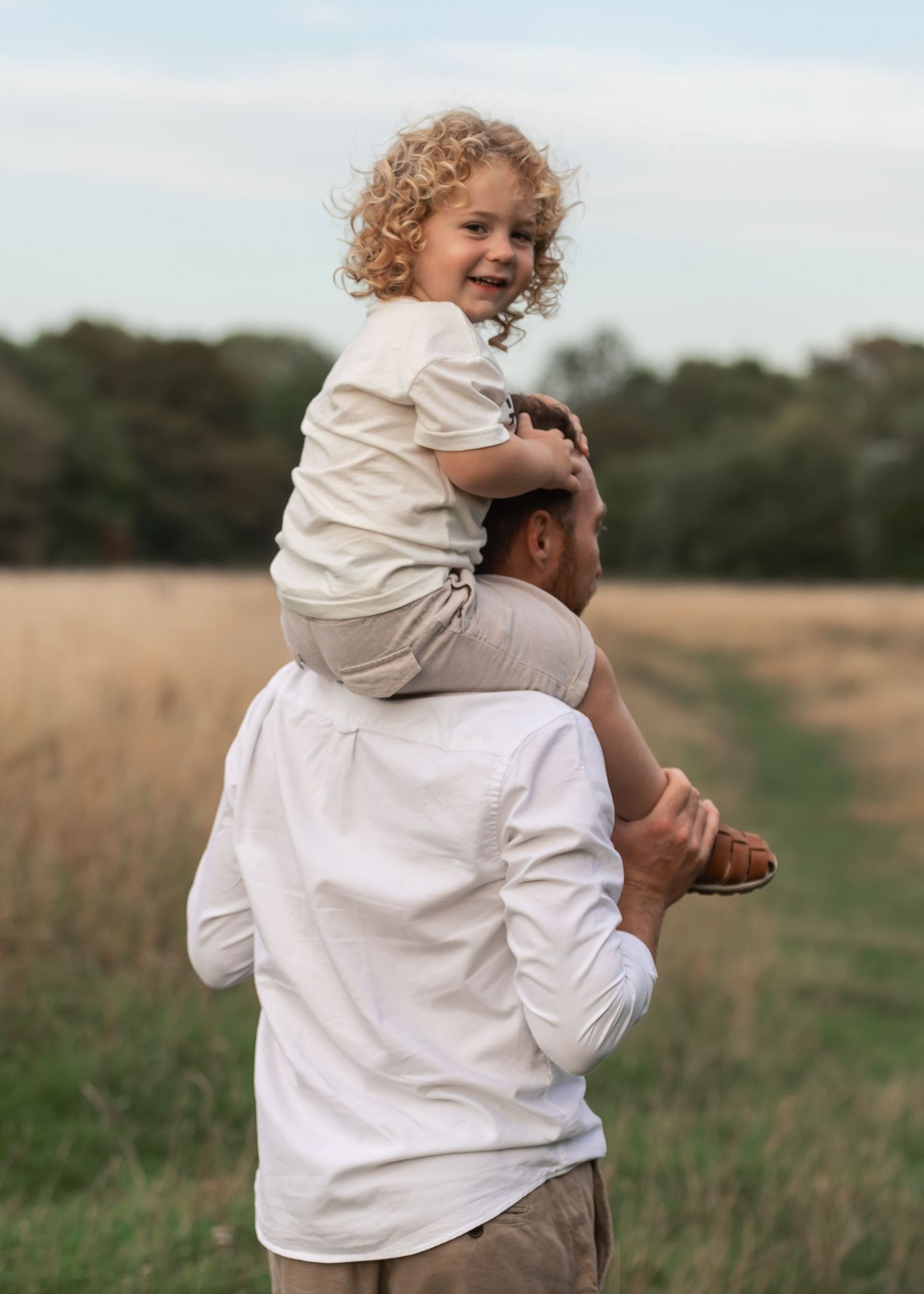 A father carries his curly-haired child on his shoulders in a sunny field, both smiling joyfully.