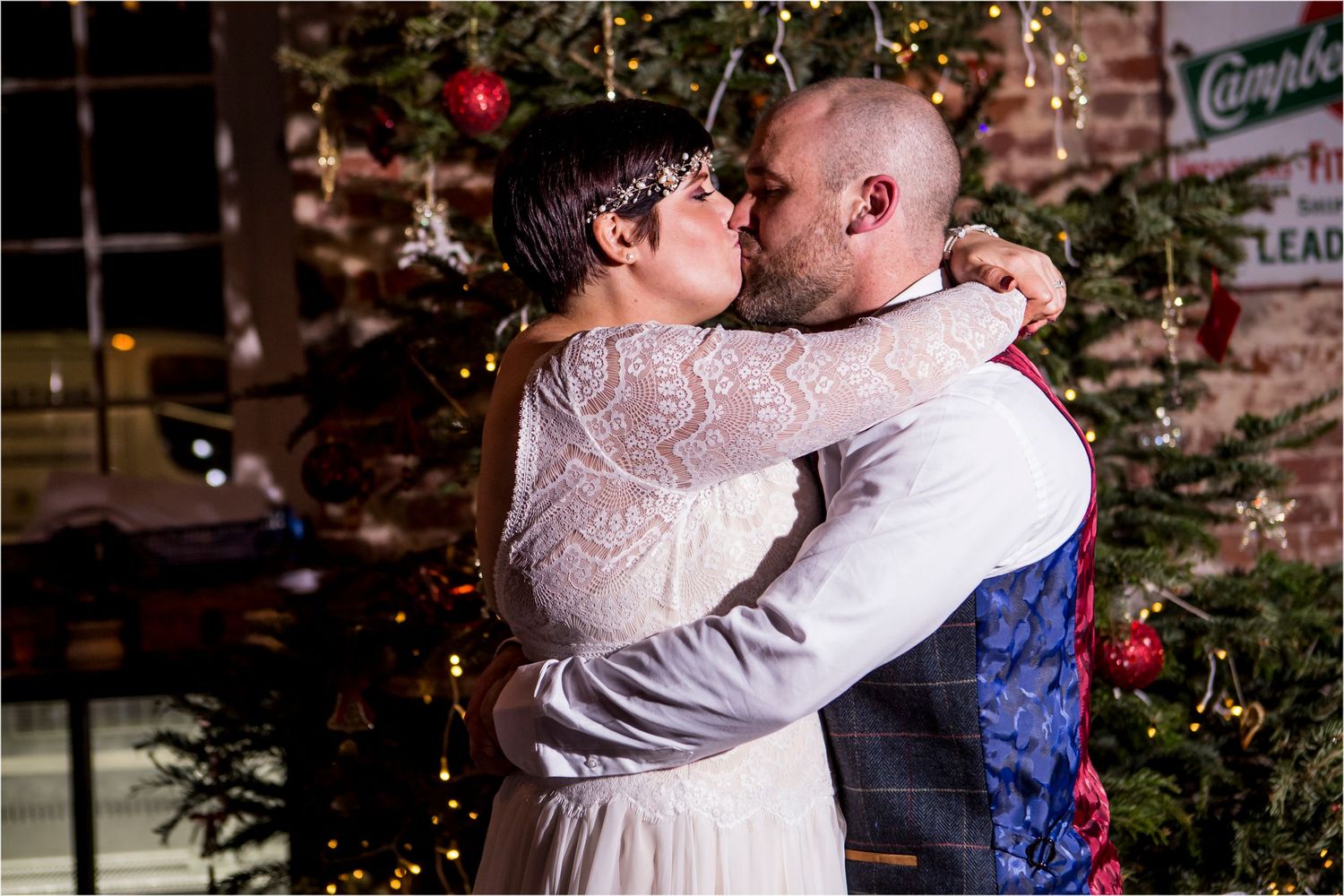 A romantic couple embraces outdoors in front of Christmas trees and twinkling holiday lights creating a magical moment.