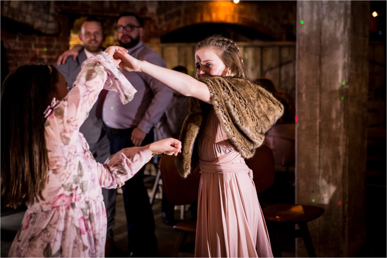 A couple dressed in vintage pink attire dances together at a dimly lit indoor venue with brick walls.