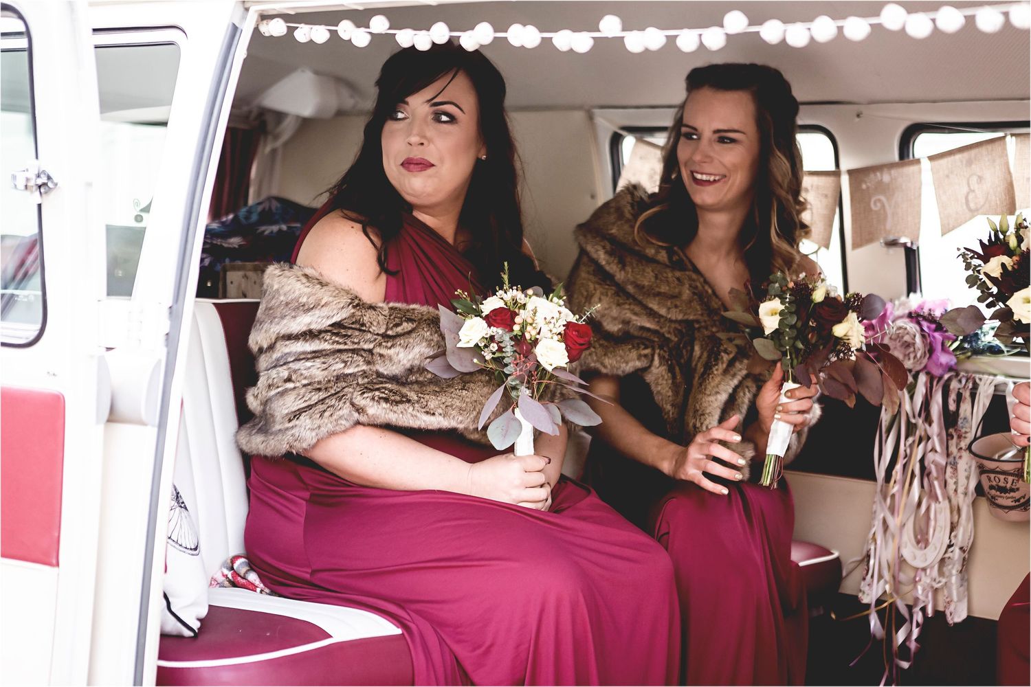 Two bridesmaids in burgundy dresses and fur wraps sit inside a vintage van decorated with string lights.