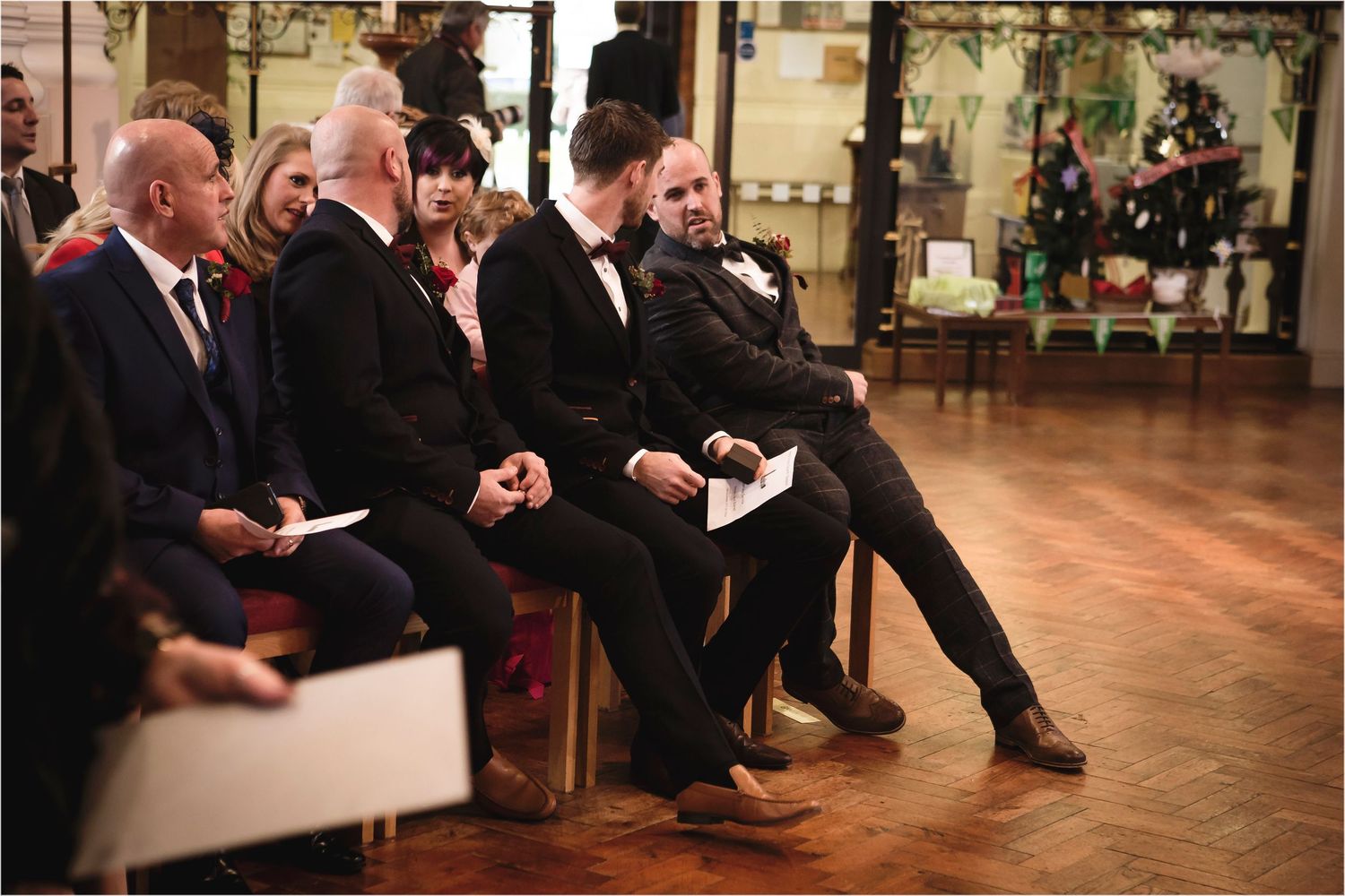 Several formally dressed attendees sit in a row on chairs during an elegant indoor event with wooden floors and decorative walls.