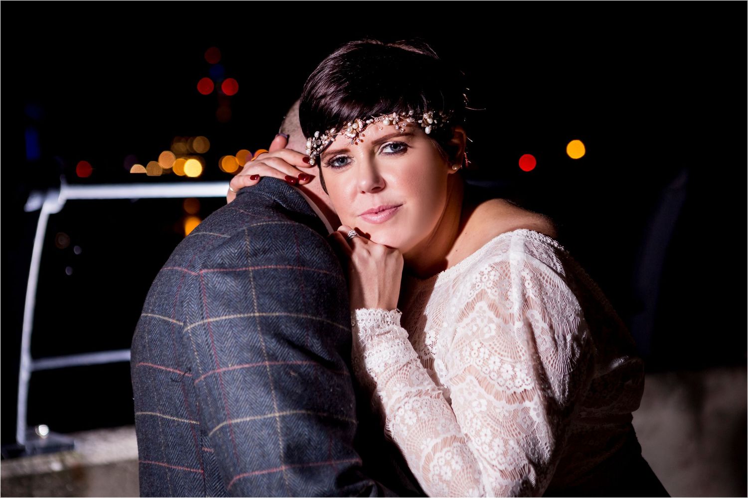 A romantic evening photo shows a couple embracing, with the person in white lace leaning on their partner against city lights.
