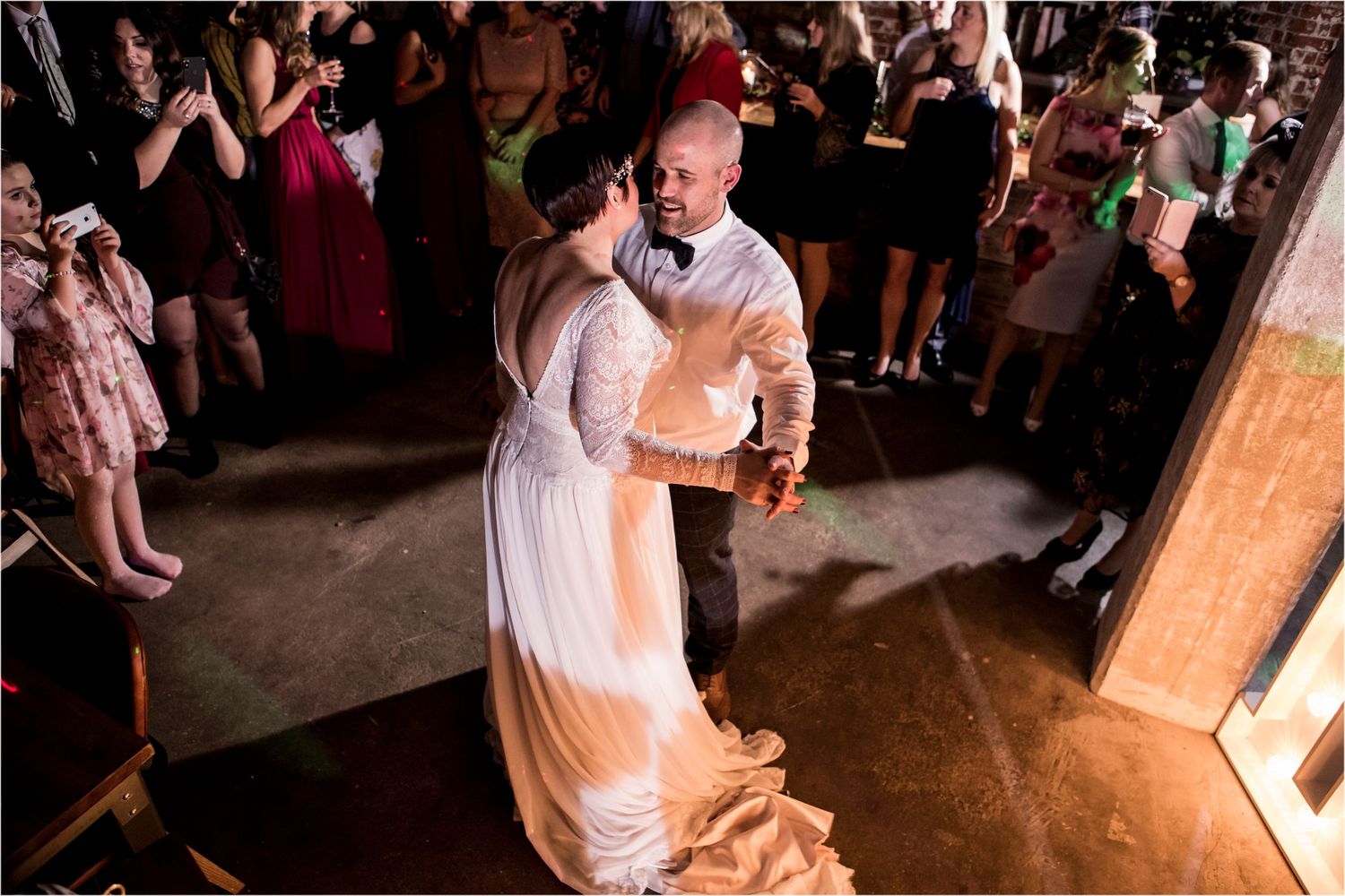 A couple shares their first dance as newlyweds on a rustic wooden dance floor surrounded by wedding guests.