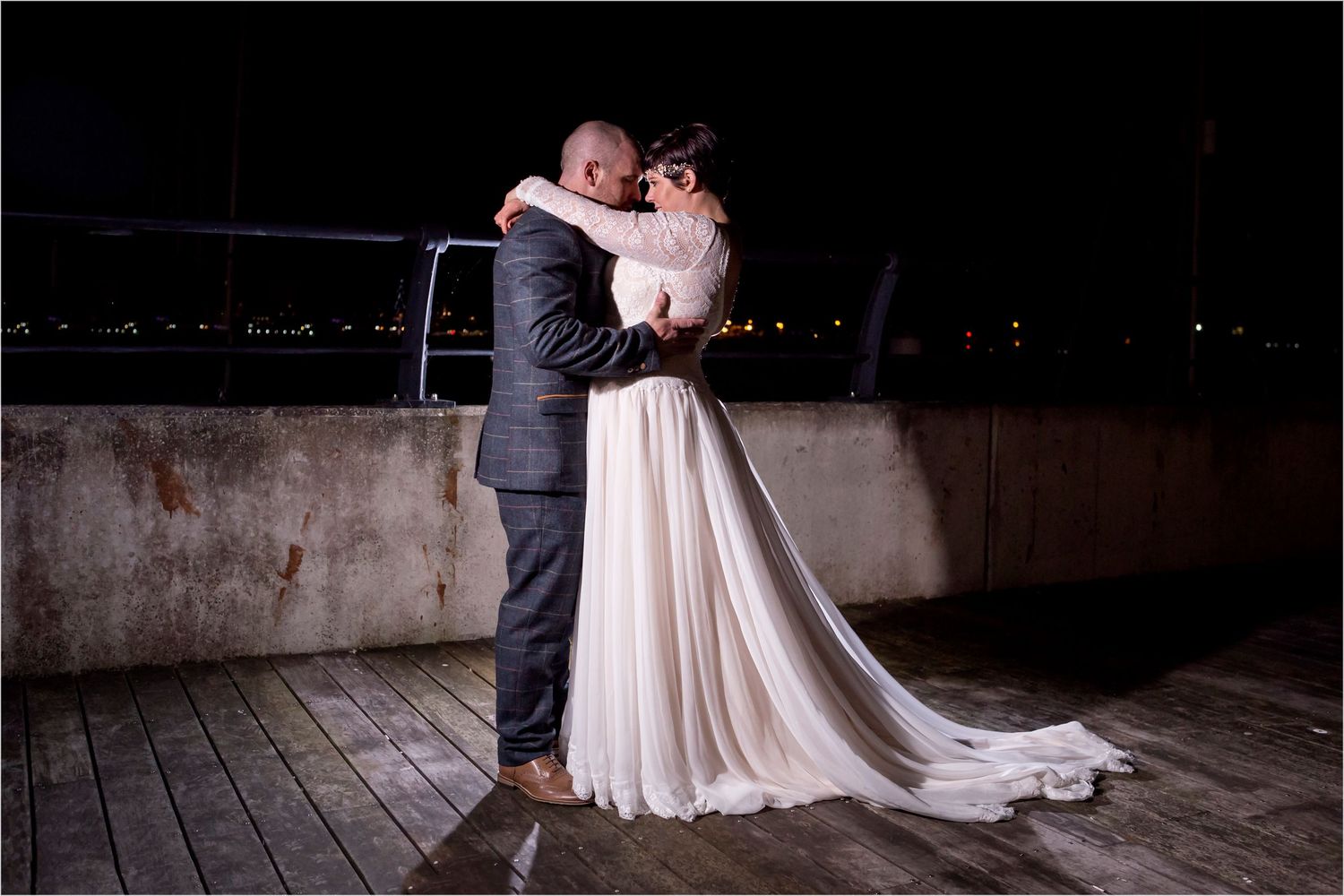 A romantic nighttime wedding scene with a couple embracing on a wooden deck with city lights glowing in the background.