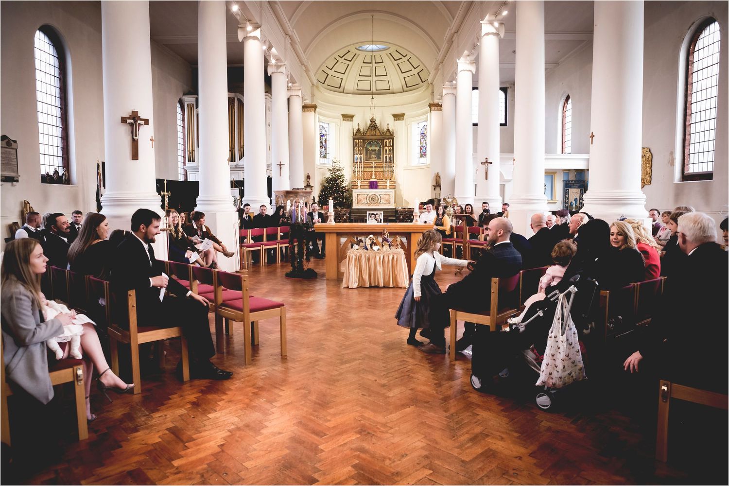 Interior of a church with guests seated in wooden pews during a ceremony on a polished herringbone floor.