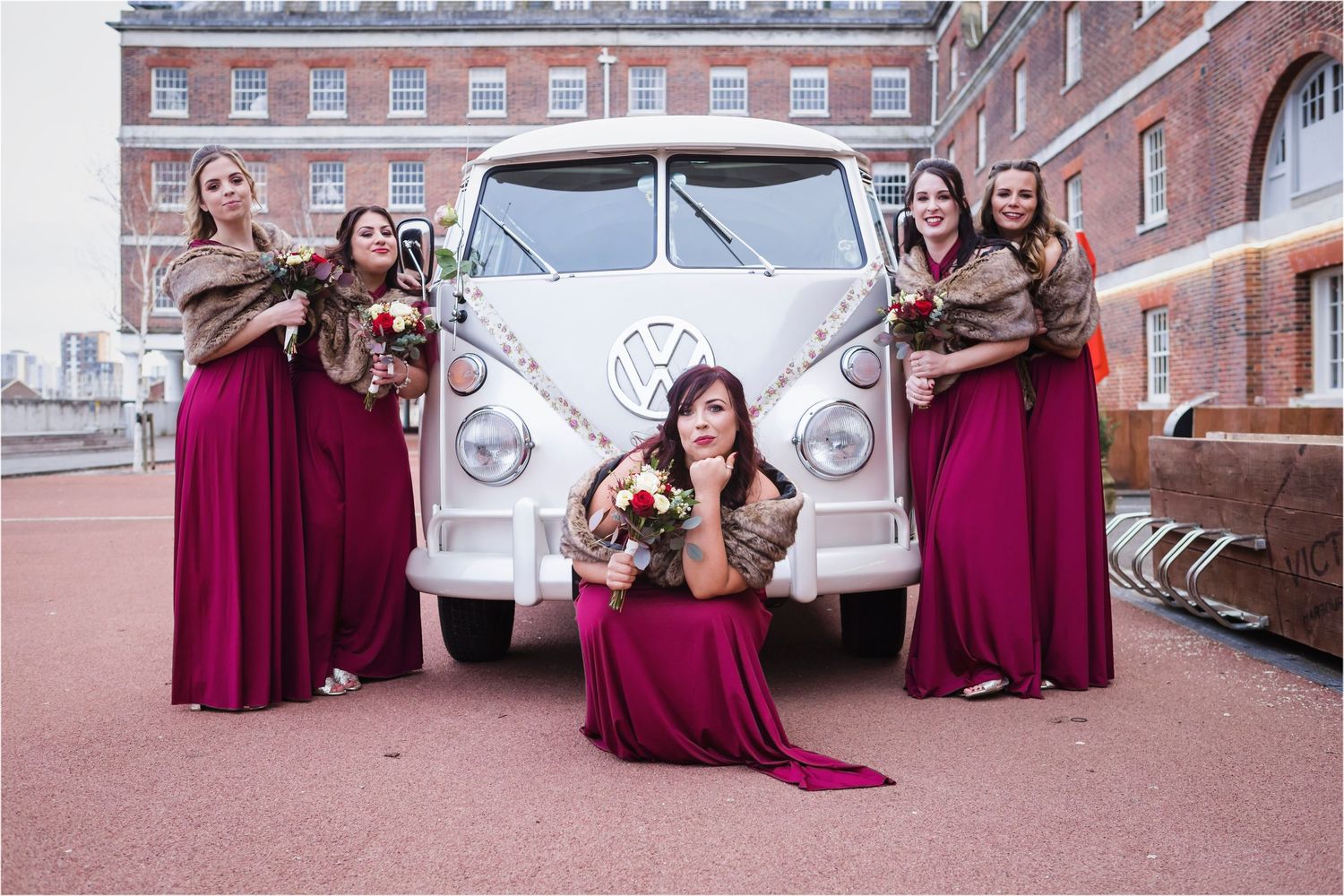 Wedding party members in burgundy dresses pose with a classic white VW bus in front of a brick building.