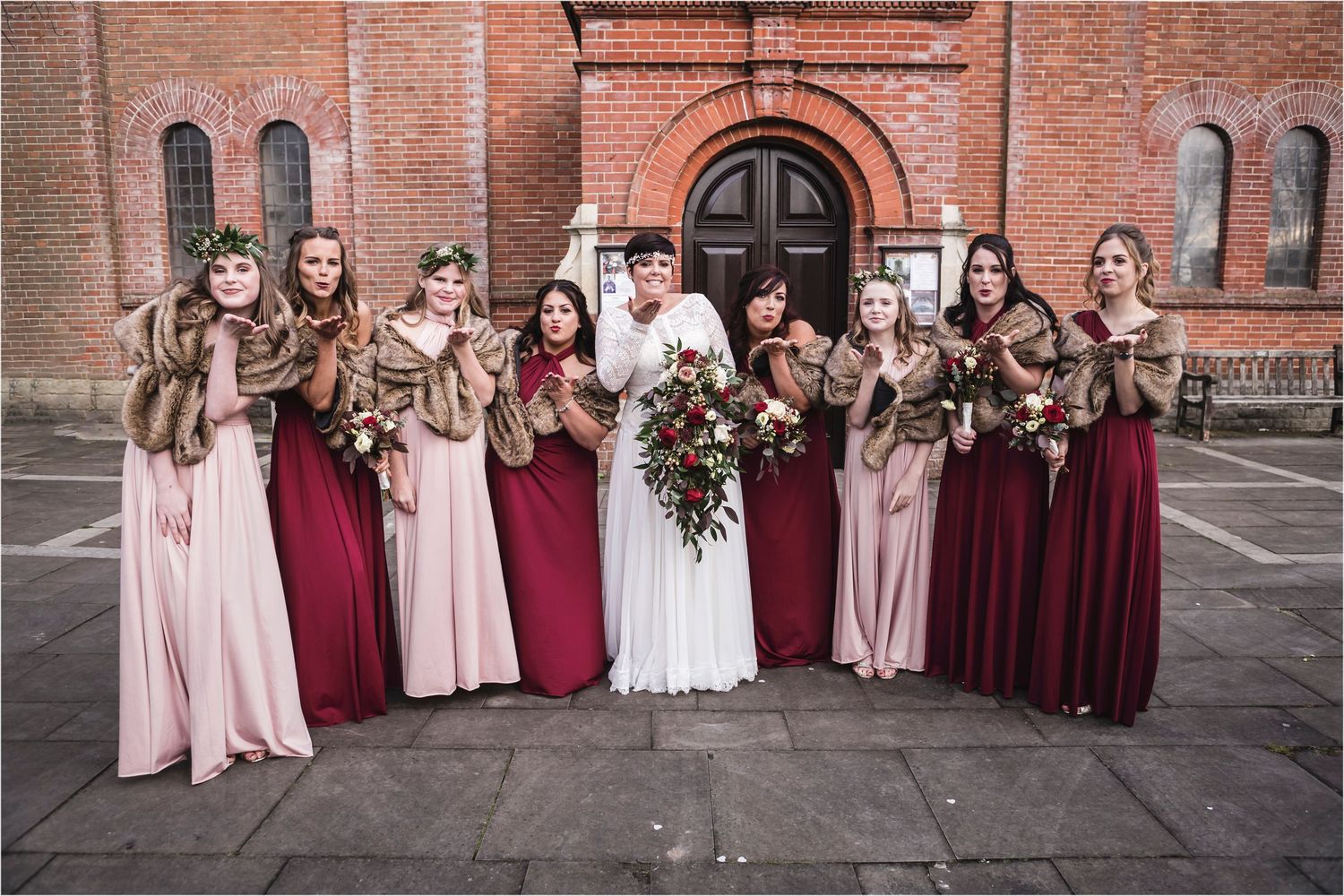 Wedding party in burgundy and blush pink dresses poses in front of historic brick church with arched doorway.