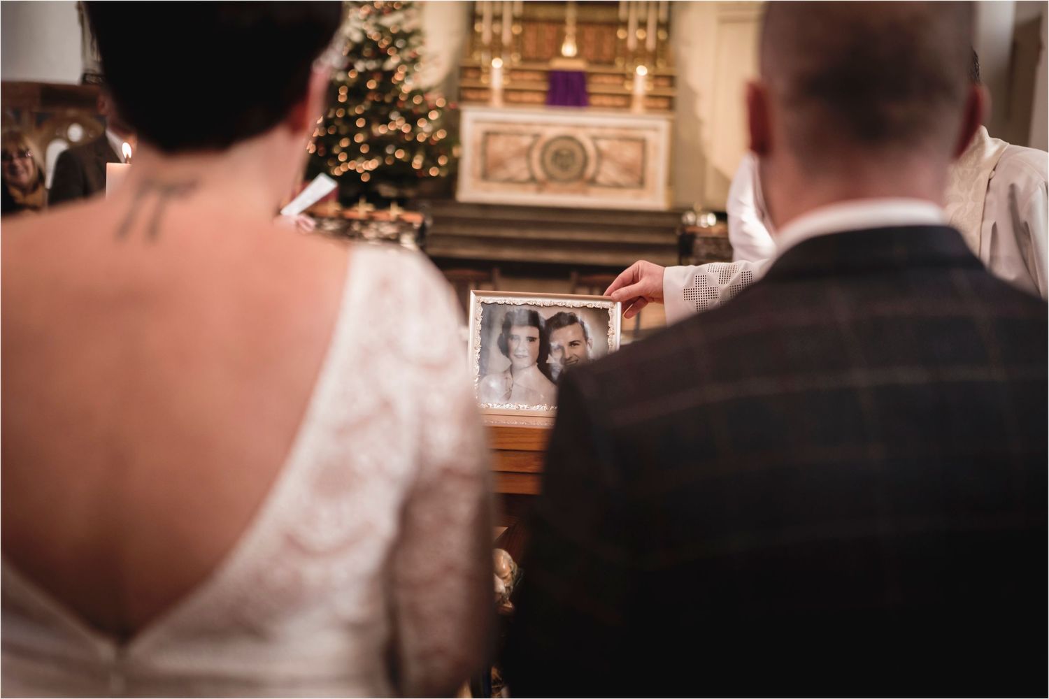 Couple sits facing forward during Christmas wedding ceremony with decorated tree and altar in background.