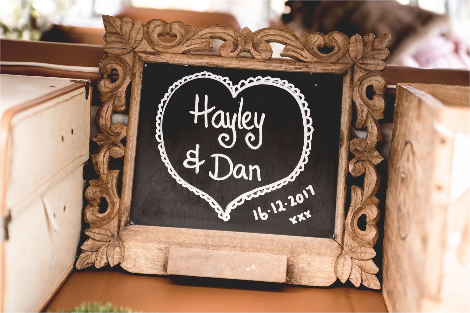 A vintage-style ornate wooden frame displays a chalkboard sign with names and a heart design in white chalk.
