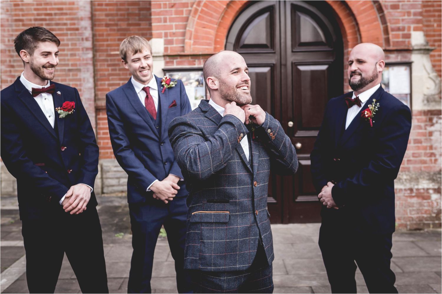 A group of groomsmen in navy suits and bow ties pose together and laugh outside a brick building.