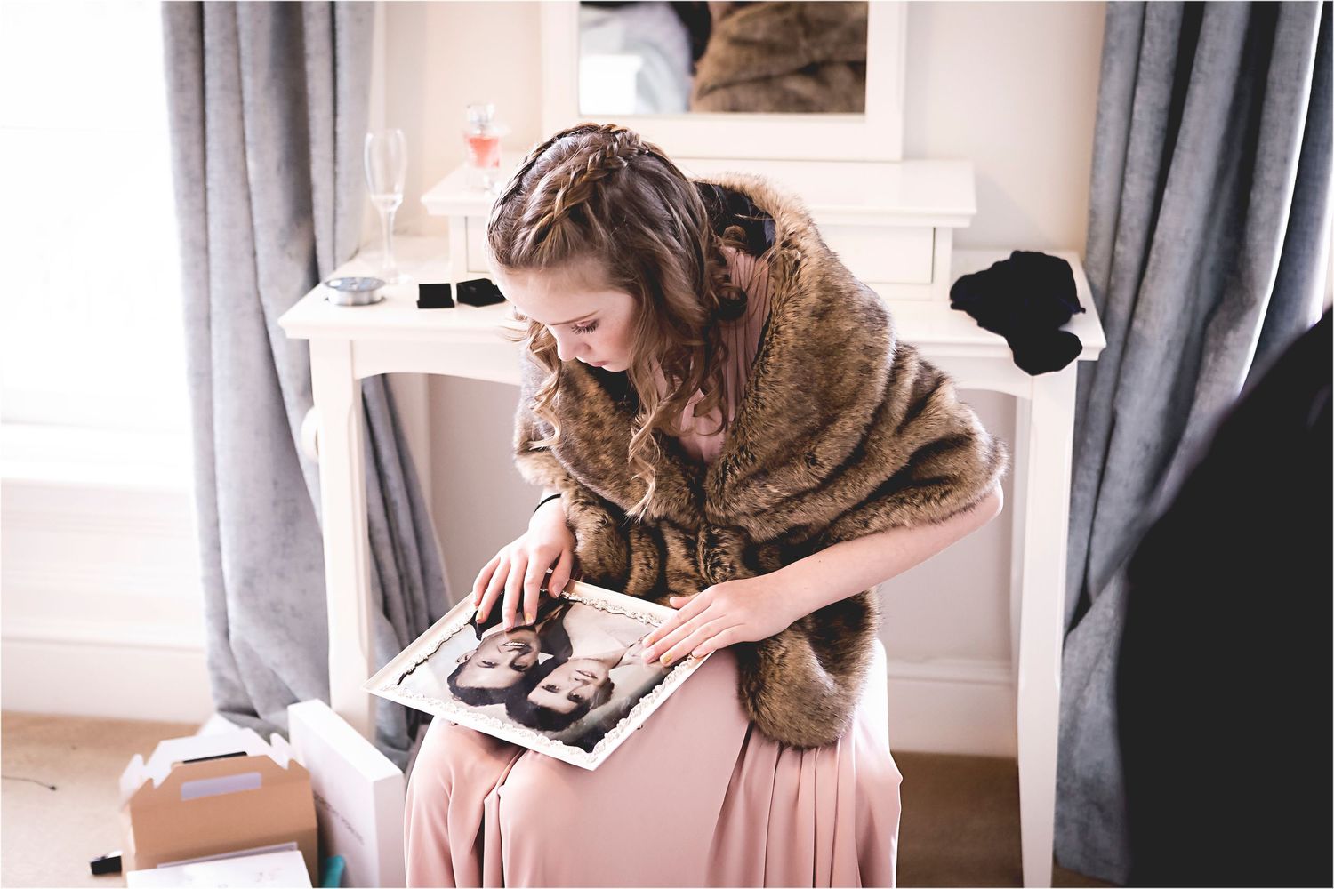 Person in brown fur coat looking down at a photo album while seated indoors near a curtained window.