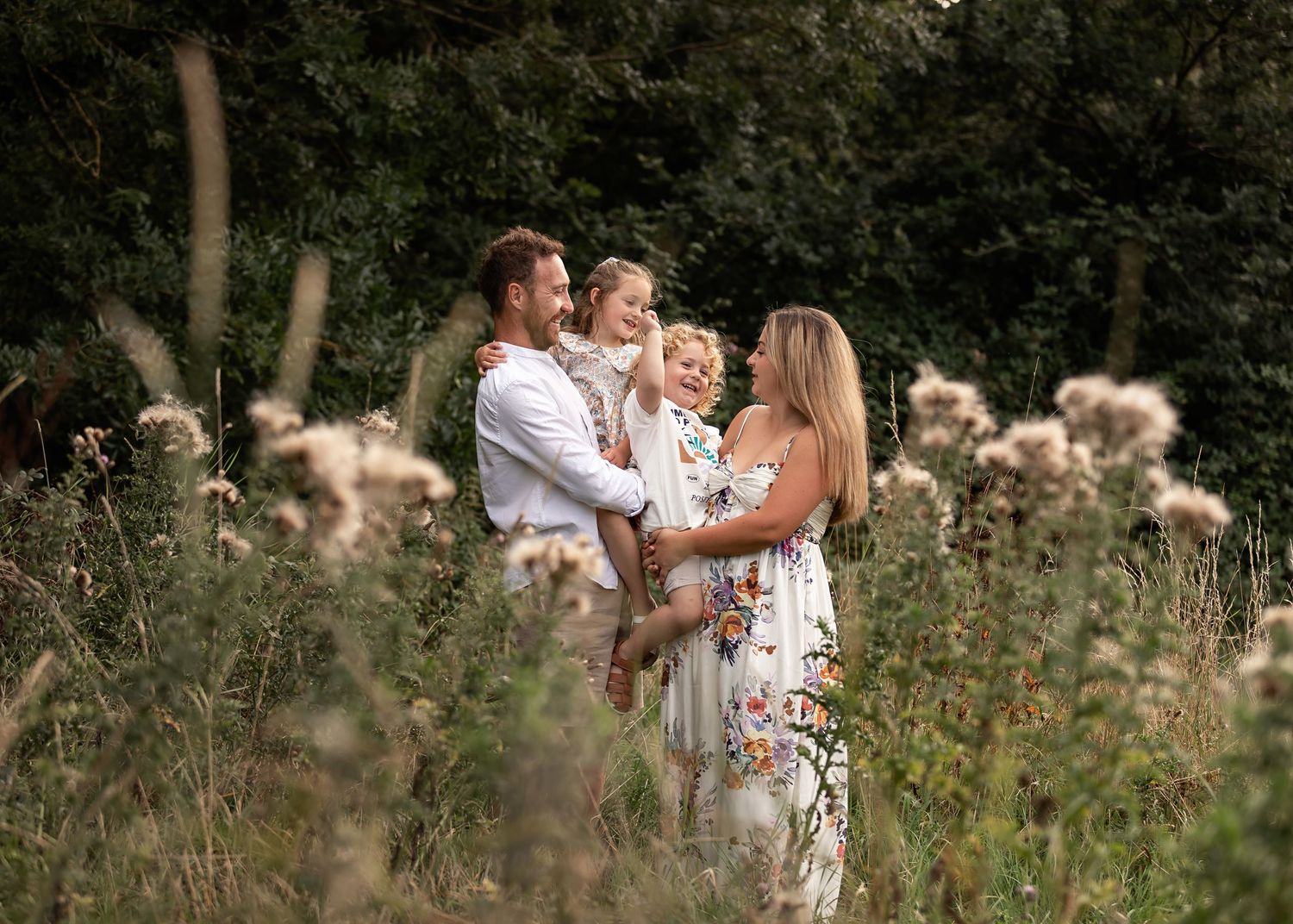 Happy family with toddler posing in a wildflower meadow surrounded by lush green trees.