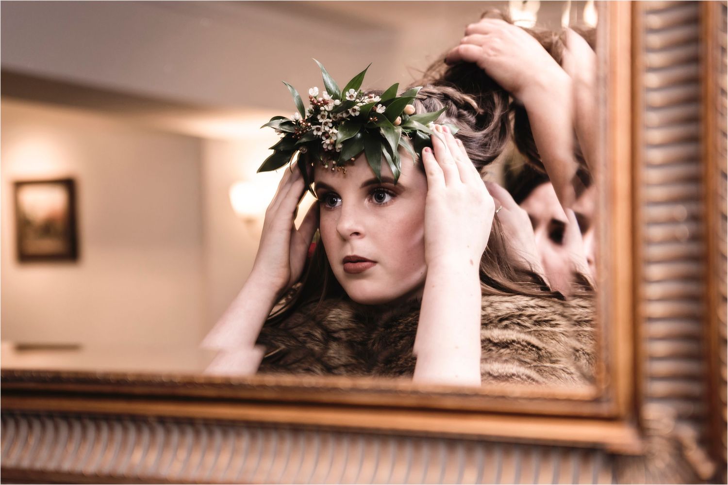Mirror reflection shows person wearing a delicate greenery crown while adjusting it in warm-toned lighting.