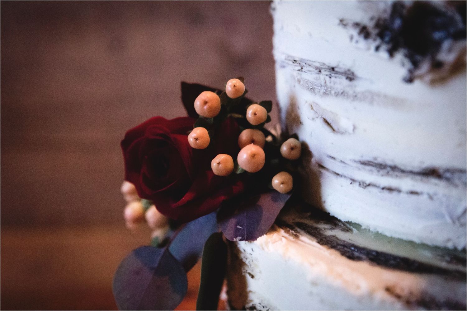 Deep burgundy roses and pink berries accent a white tiered wedding cake in a moody, romantic setting.