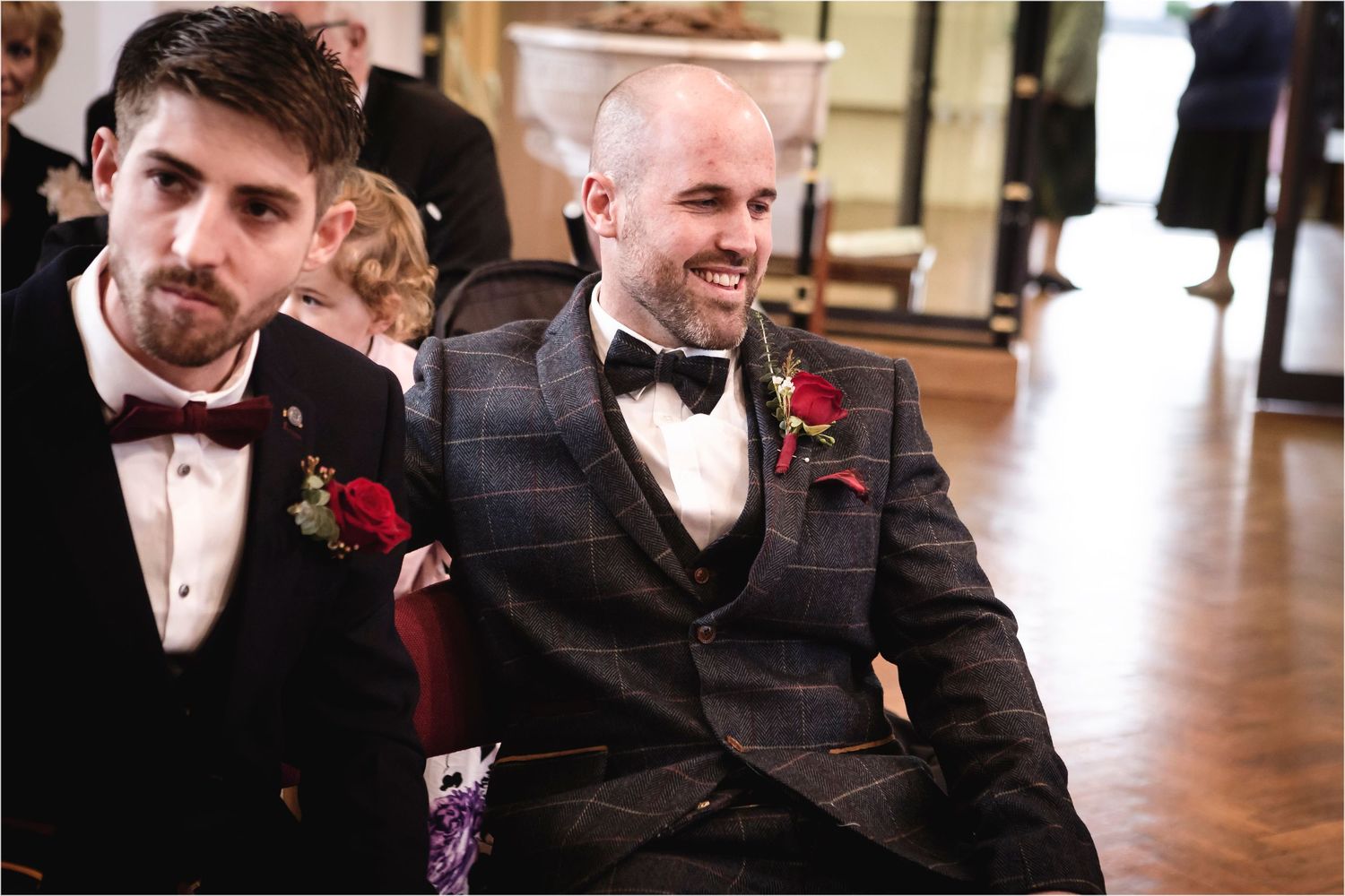 Two wedding party members in black tuxedos and bow ties with red boutonnieres share a joyful moment on a wooden dance floor.