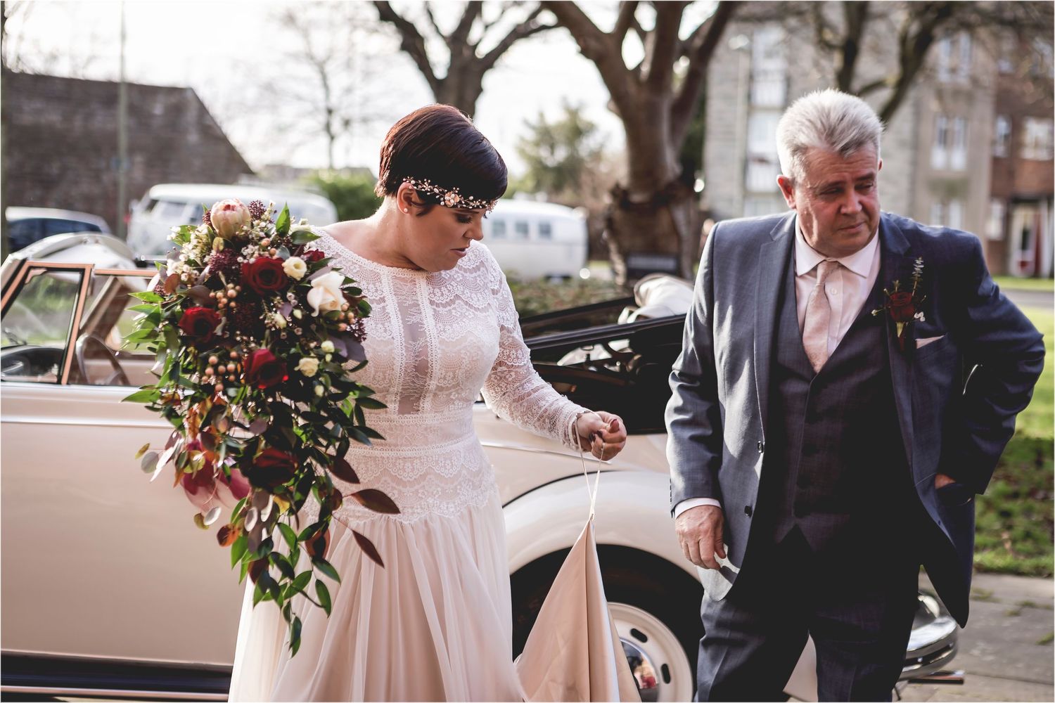 A person in a wedding dress accepts assistance from someone in a dark suit while exiting a vintage pink car with flowers.