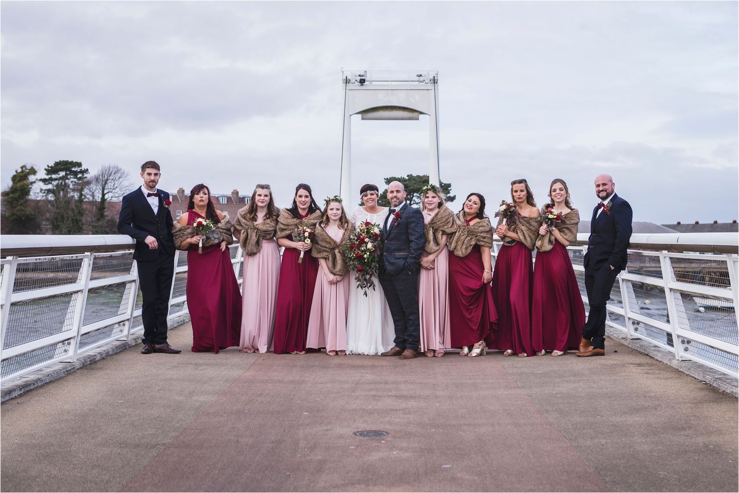 Wedding party in burgundy bridesmaid dresses poses on white bridge during cloudy day.