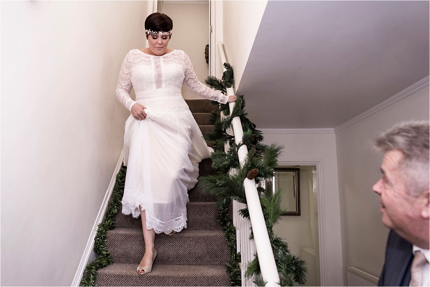 A person in a white lace dress descends a staircase decorated with greenery and white flowers.
