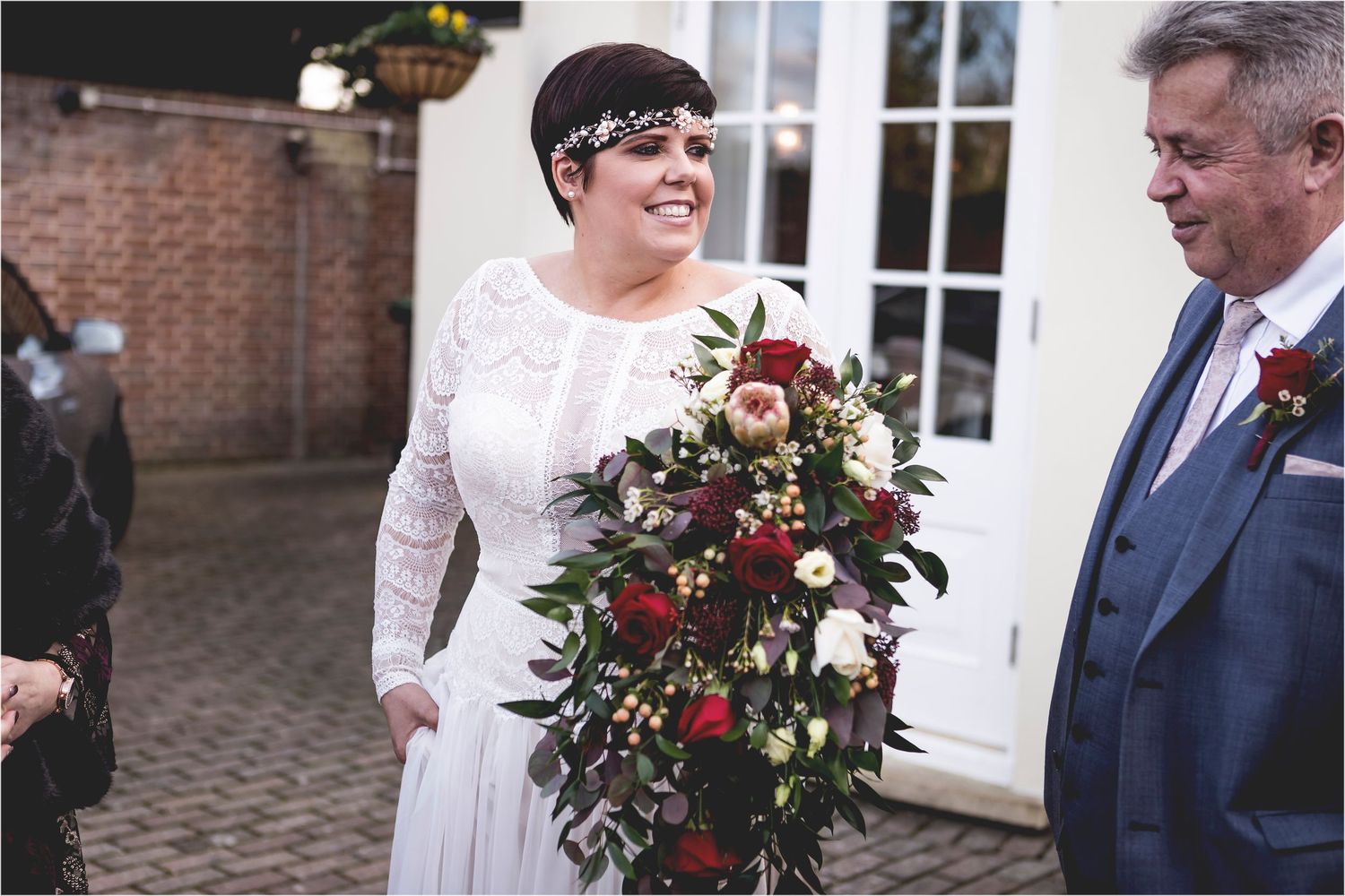 A joyful bride in a long-sleeved lace gown holds a cascading burgundy and blush floral bouquet at an outdoor venue.