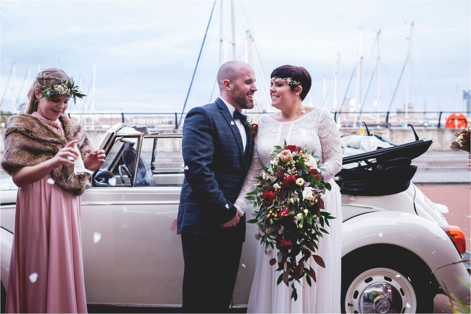 A wedding couple shares a moment near boats at a marina while another guest watches in a pink dress.