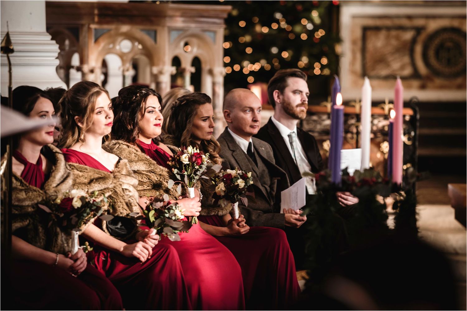 Wedding party in formal attire sits in church pews with red dresses and candlelit decor during holiday celebration.