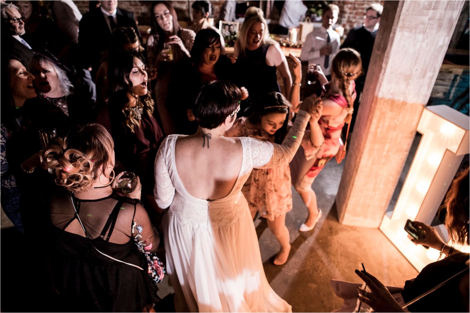 A crowded dance floor at a wedding reception shows people dancing and celebrating in a dimly lit venue.