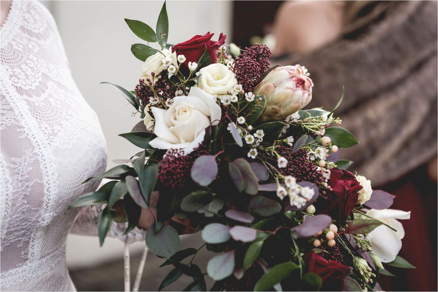 A wedding bouquet featuring burgundy and white roses with eucalyptus leaves and dark berries against a lace backdrop.