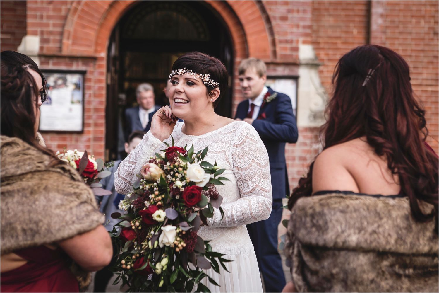 A joyful bride in a lace dress holds a deep red and burgundy floral bouquet while standing outside a brick building.