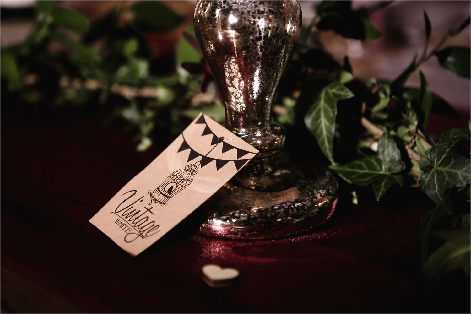 A wine glass with a decorative tag featuring bunting flags and drawings sits among green foliage in moody lighting.
