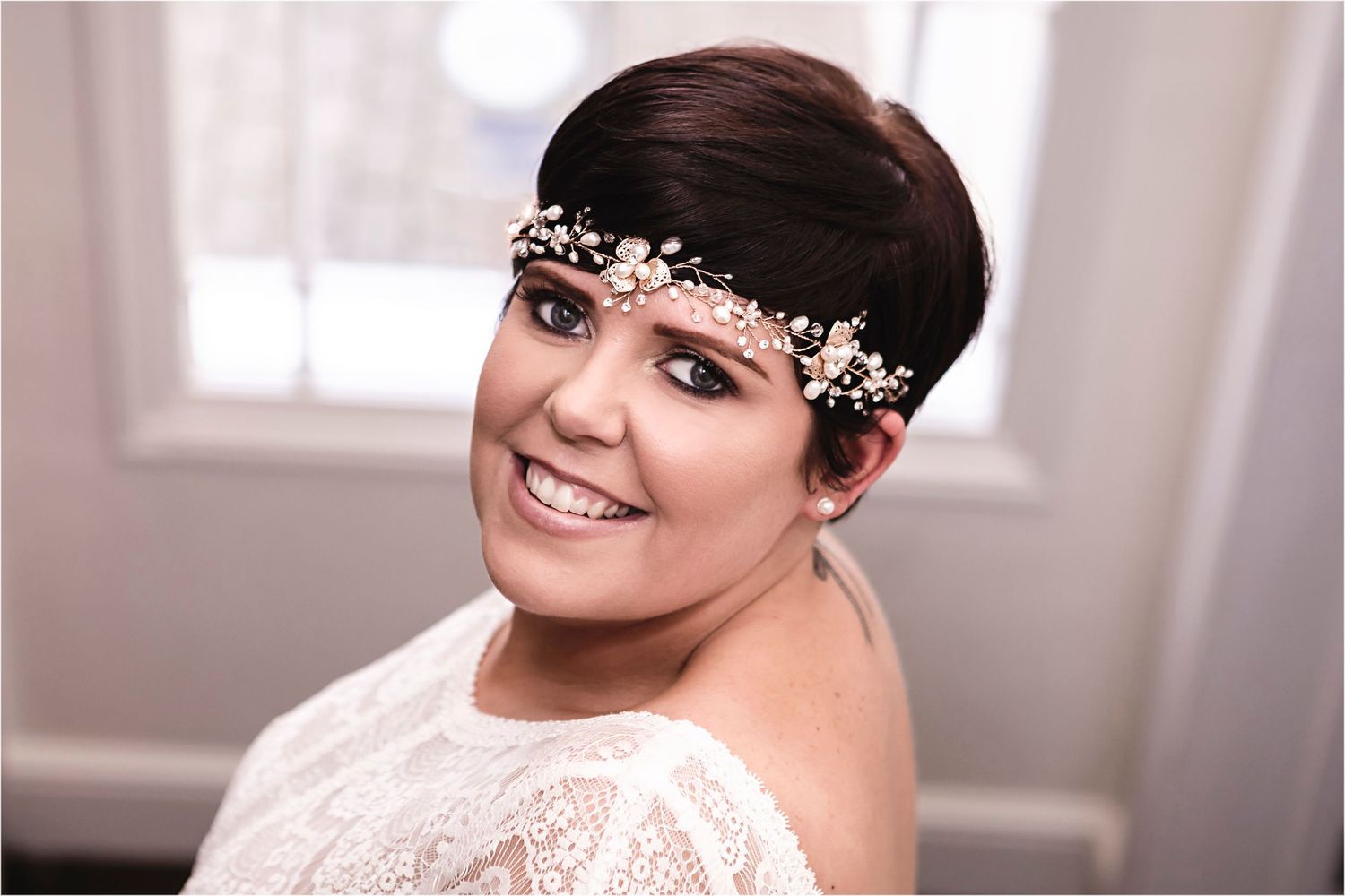 Smiling bride wearing delicate floral crystal headpiece with white lace dress in soft window light.