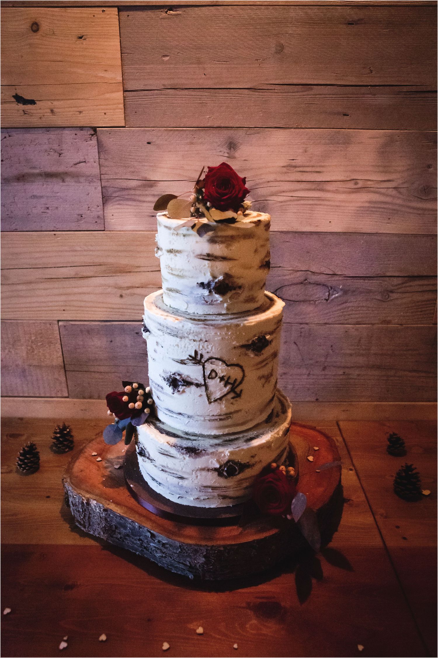 Rustic three-tier birch bark wedding cake with burgundy roses displayed on wooden slab with pinecone decorations.