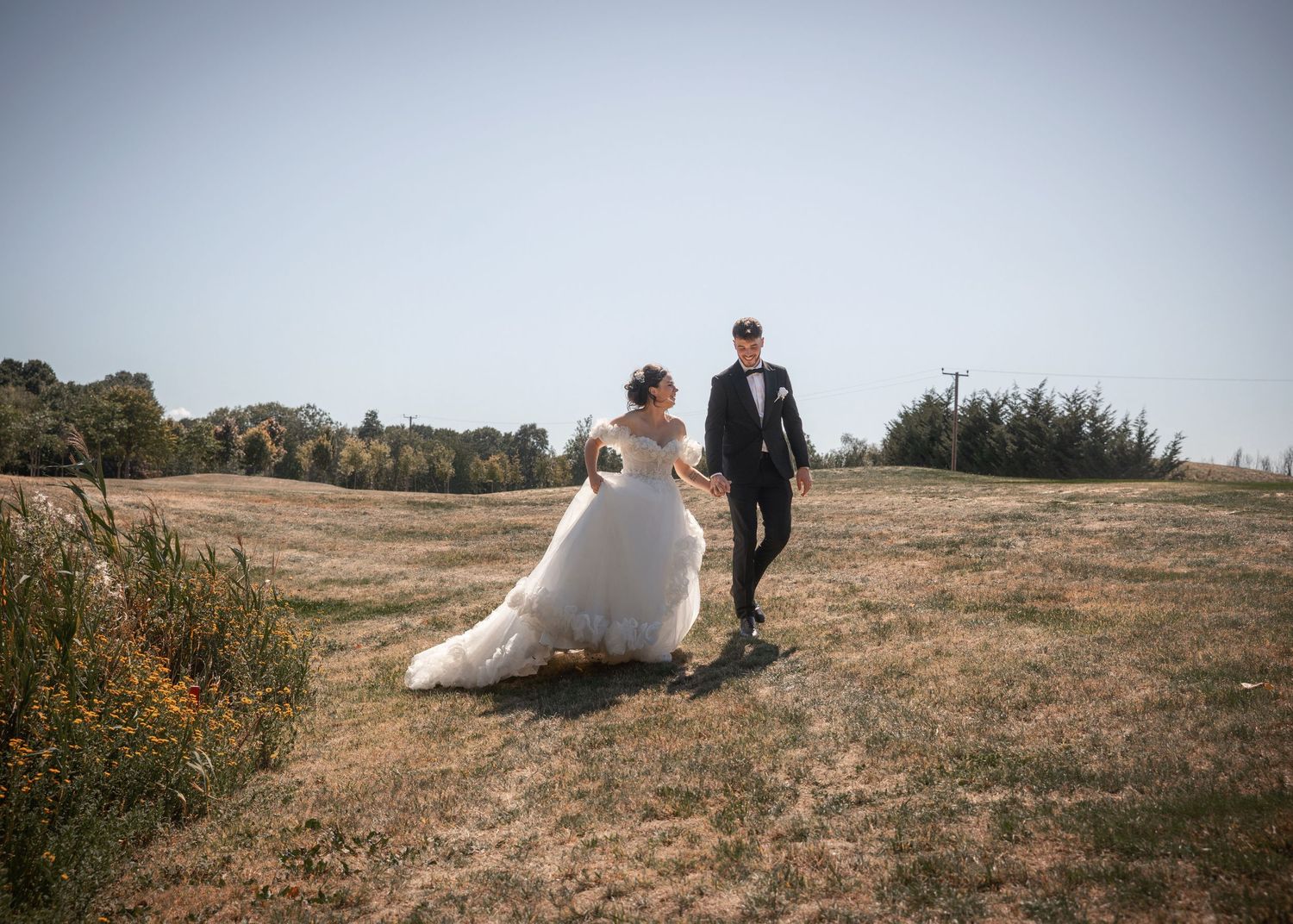 Couple in formal wedding attire sits on grassy hillside overlooking scenic landscape at sunset.