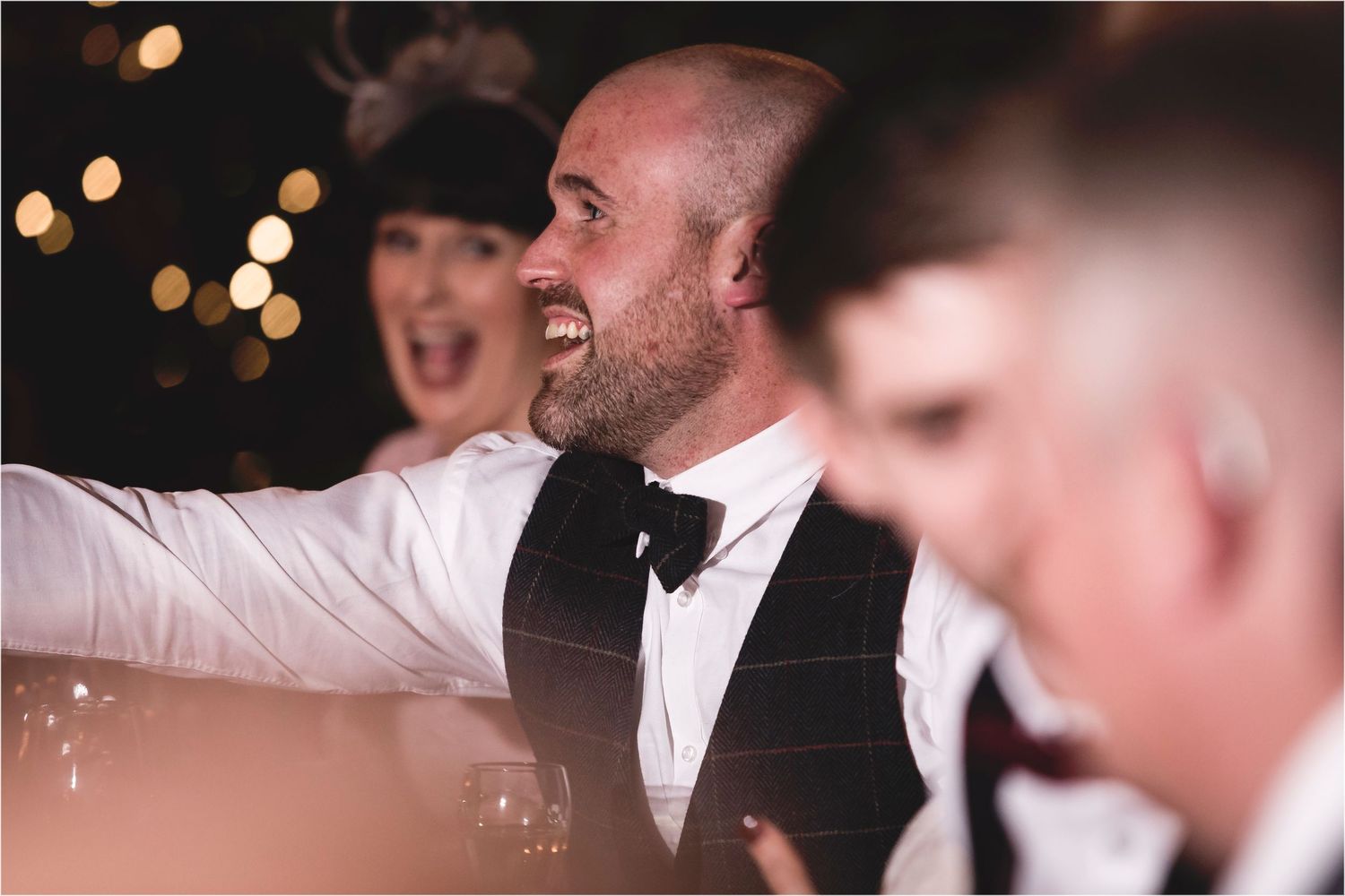 A joyful guest in formal attire laughs during a wedding reception with bokeh lights in the background.