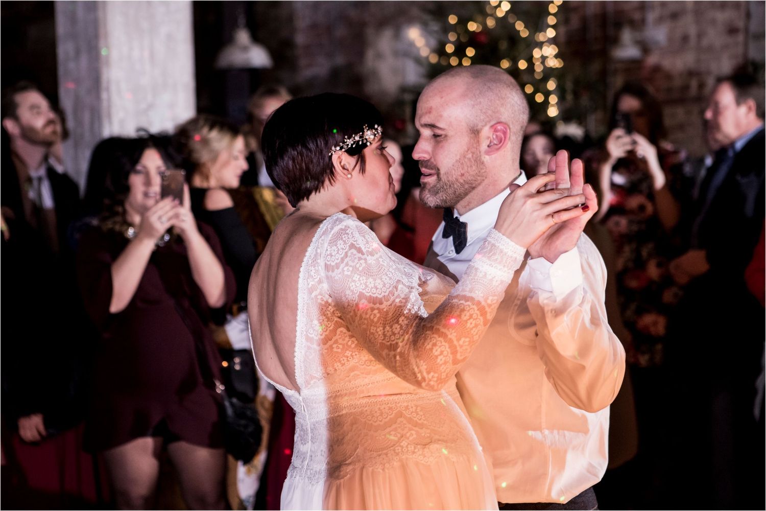 A couple in wedding attire share a romantic dance together at a reception with guests visible in the background.