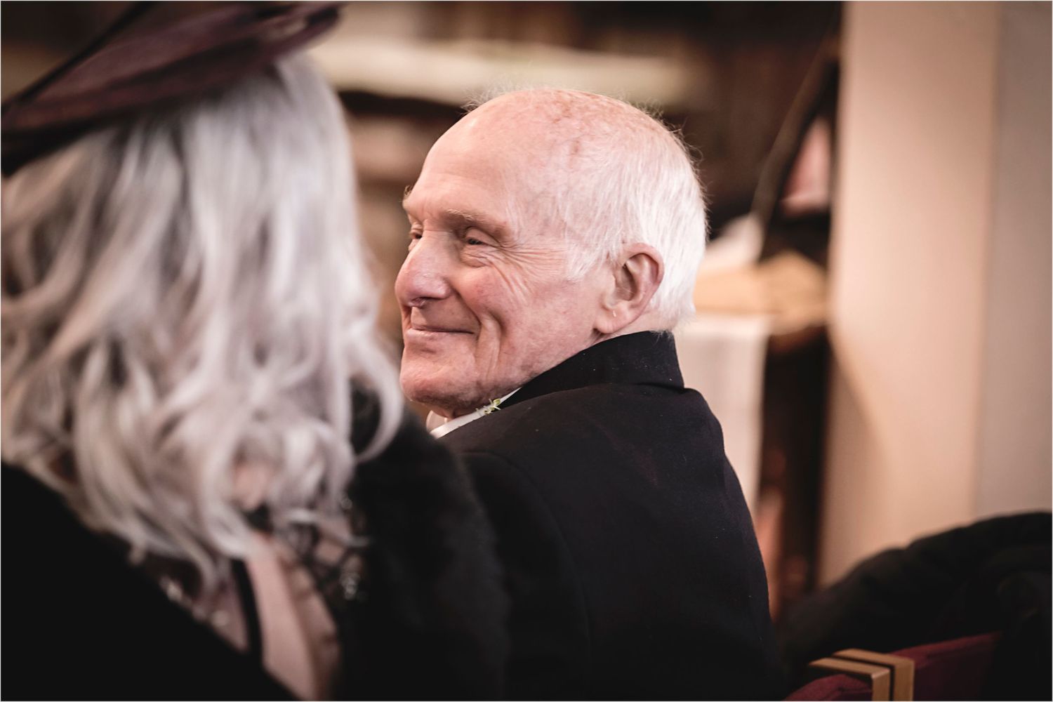 An elderly person in a black coat smiles warmly while engaging in conversation.