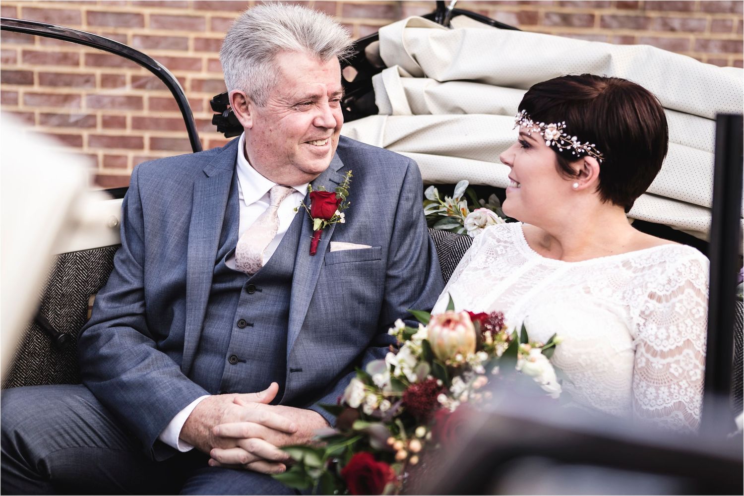Two wedding guests share a joyful moment together while seated in an elegant carriage with brick backdrop and floral decor.
