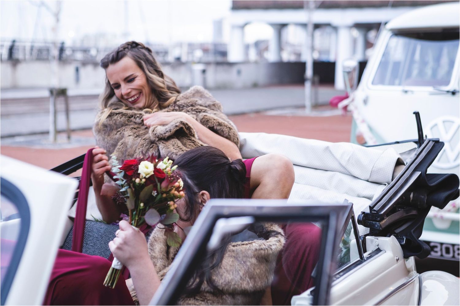 Someone leaning out of a vintage car joyfully holding a flower bouquet while wearing a furry coat on a cloudy day.