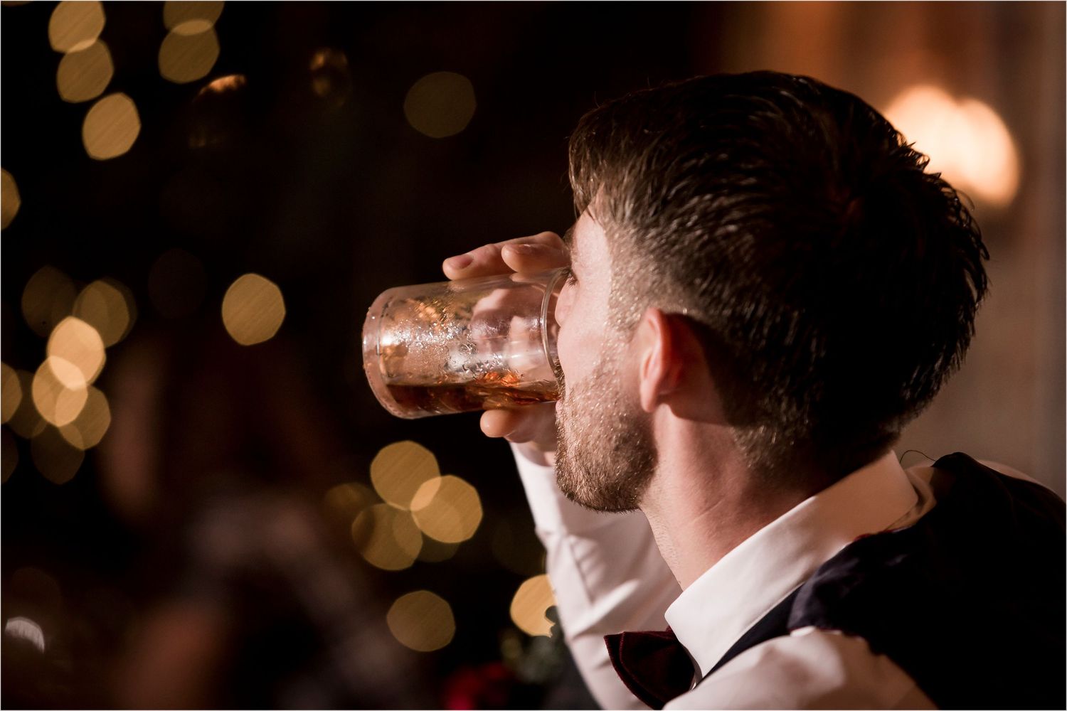 A person in formal attire drinks from a glass with warm glowing bokeh lights in the background.