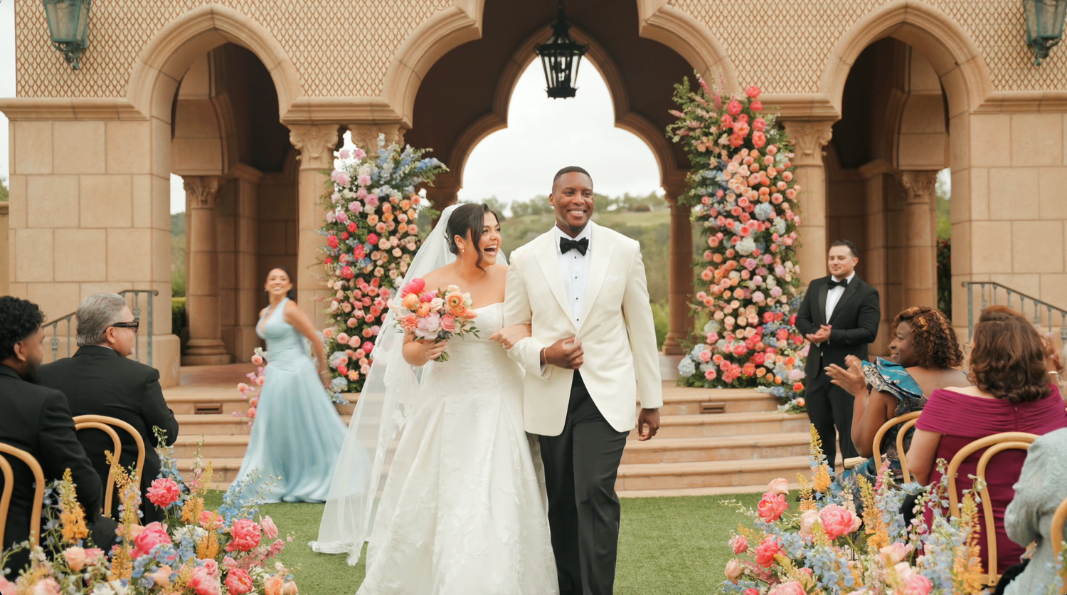 Bride and groom walking down floral aisle at outdoor wedding ceremony with colorful pink flower arrangements.
