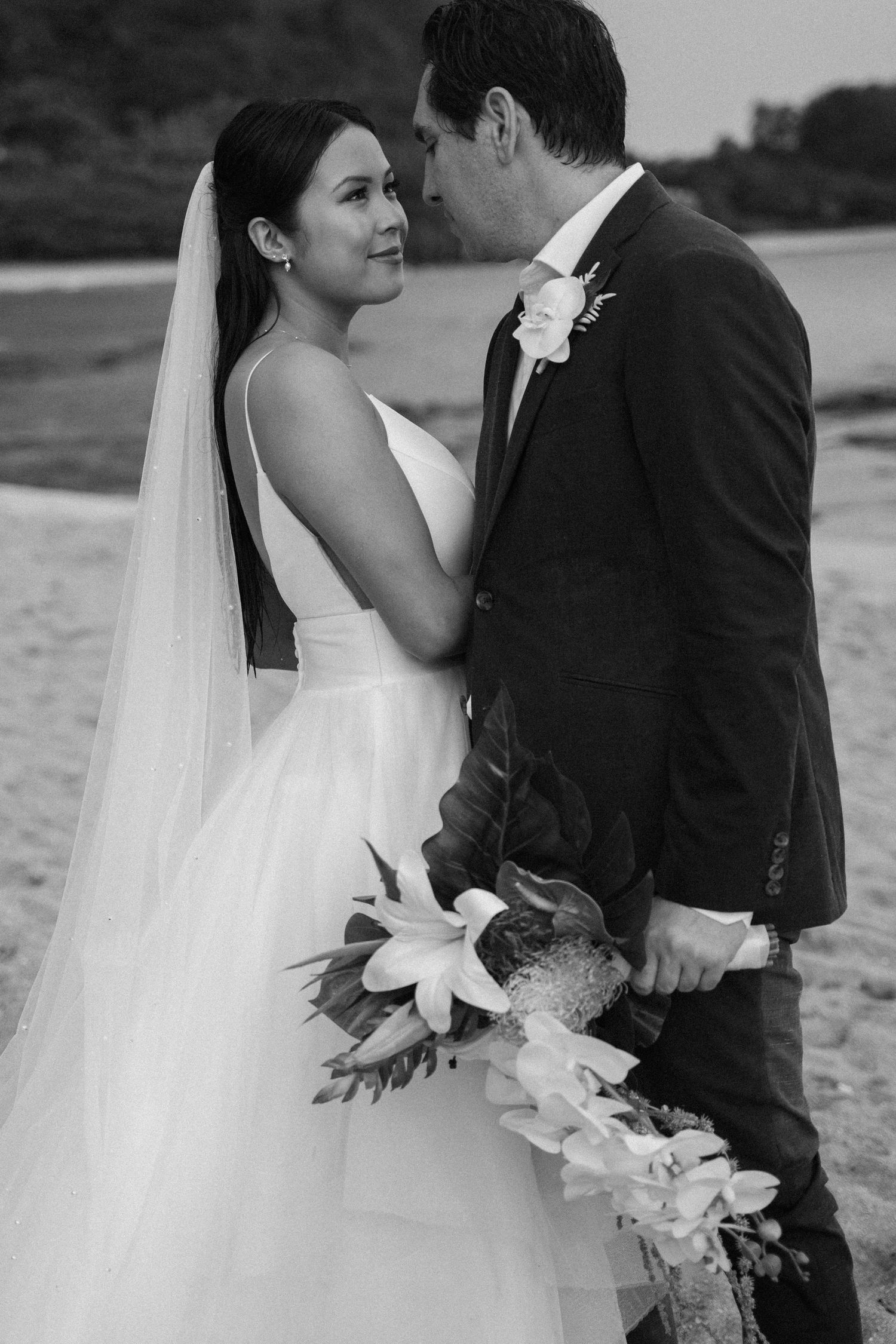 Black and white wedding photo of bride in white gown with veil holding bouquet, gazing at groom in suit on beach.