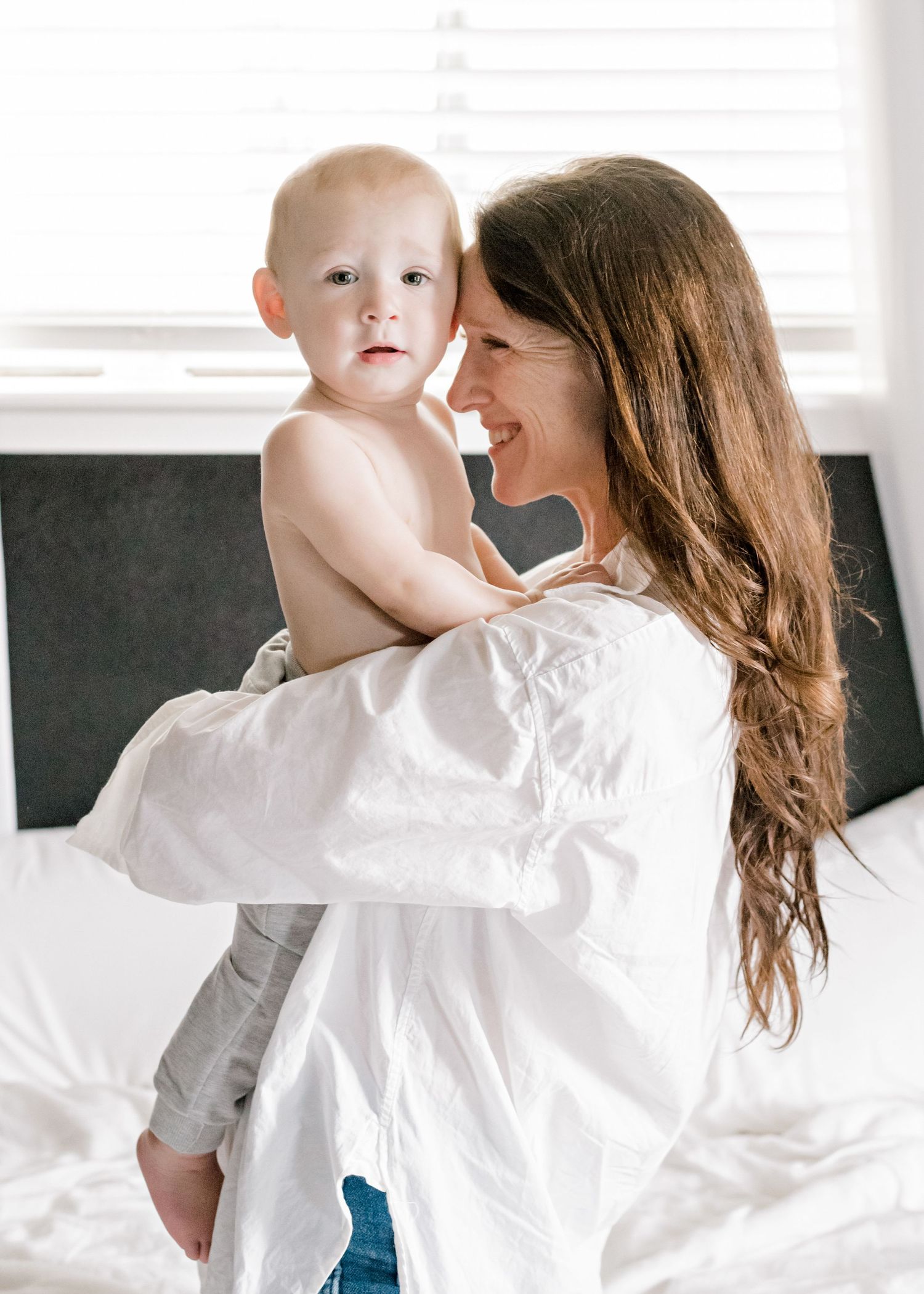 Austin, TX mom kneeling on her bed while holding her baby boy. her forehead is on his cheek as he looks towards the came