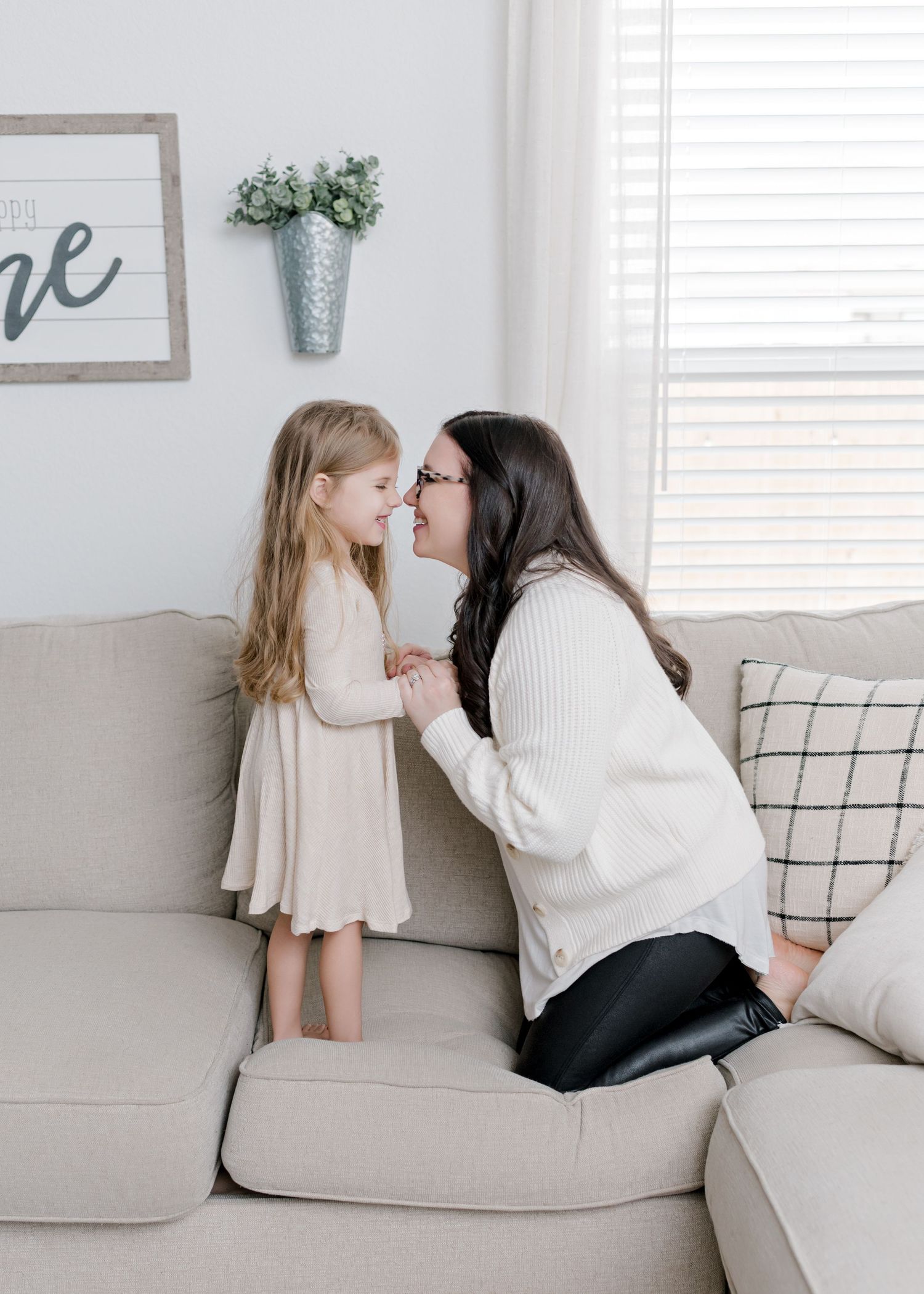 mom kneeling on the couch as her daughter stands in front of her as they touch noses and smile